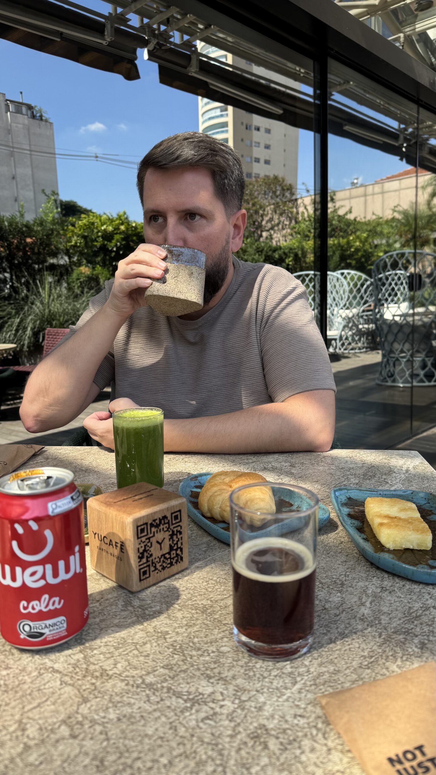 Rudi bebendo cappuccino na área externa do YUCAFÉ dentro do Pullman Ibirapuera com suco verde e toast na mesa