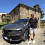 Rudi ao lado do Alfa Romeo cinza na frente da hospedagem de pedra na Toscana