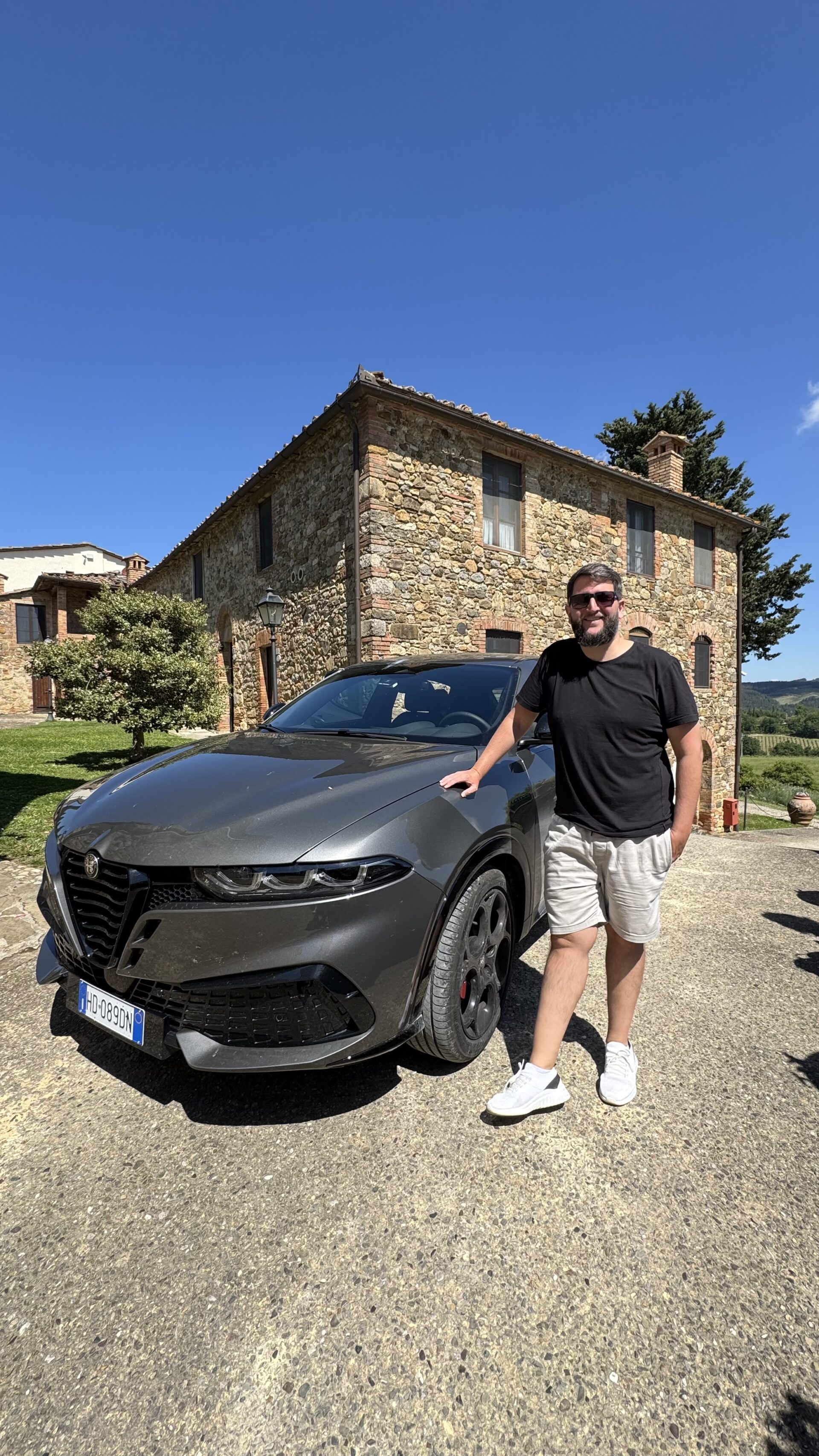 Rudi ao lado do Alfa Romeo cinza na frente da hospedagem de pedra na Toscana