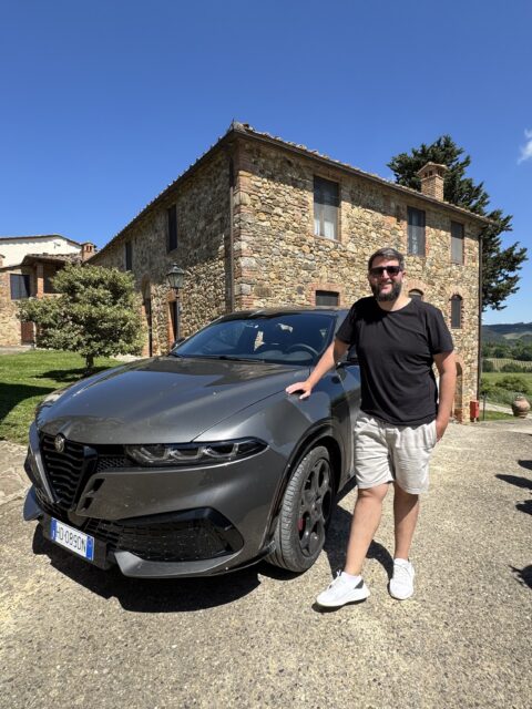 Rudi ao lado do Alfa Romeo cinza na frente da hospedagem de pedra na Toscana