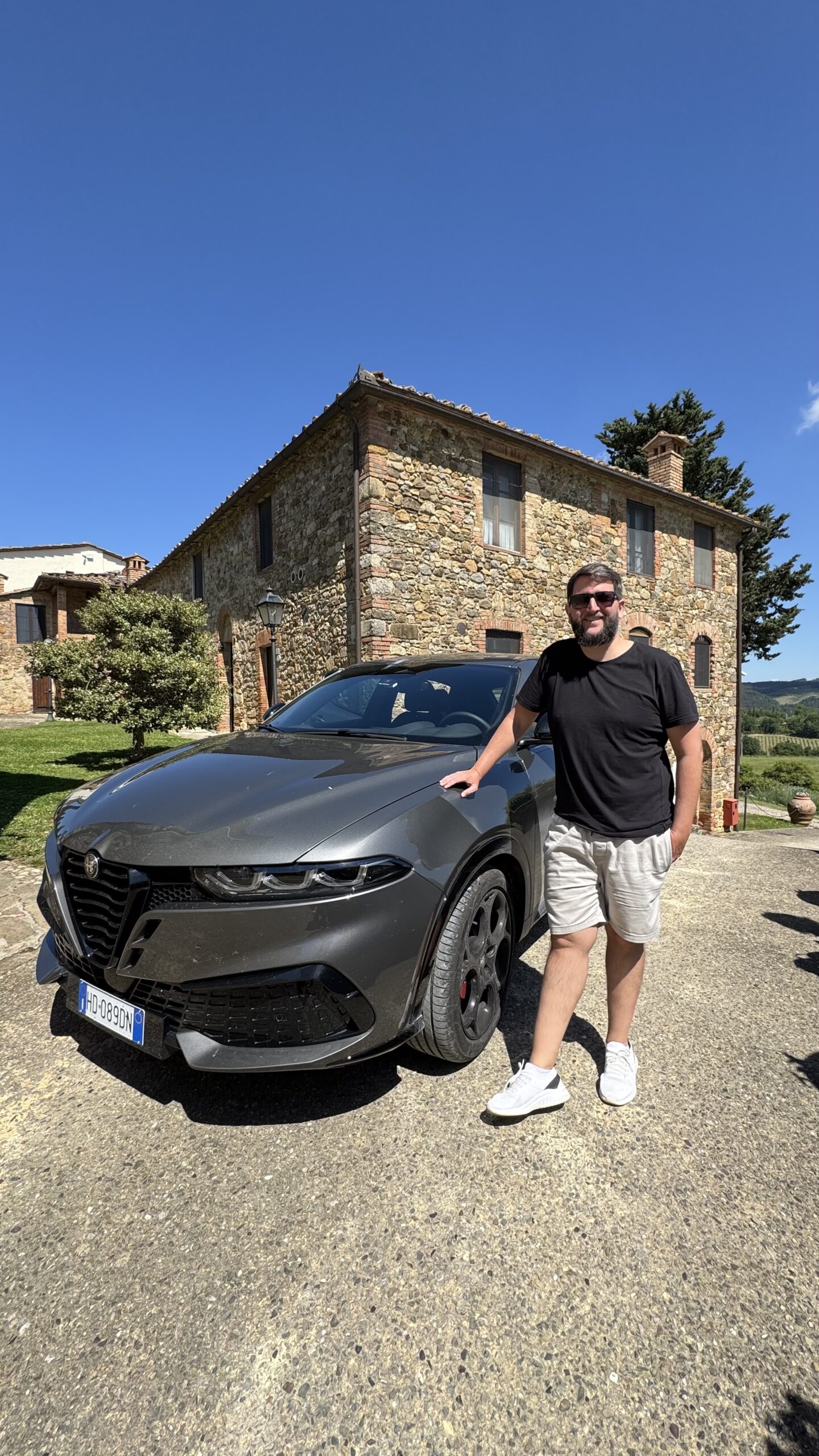Rudi ao lado do Alfa Romeo cinza na frente da hospedagem de pedra na Toscana