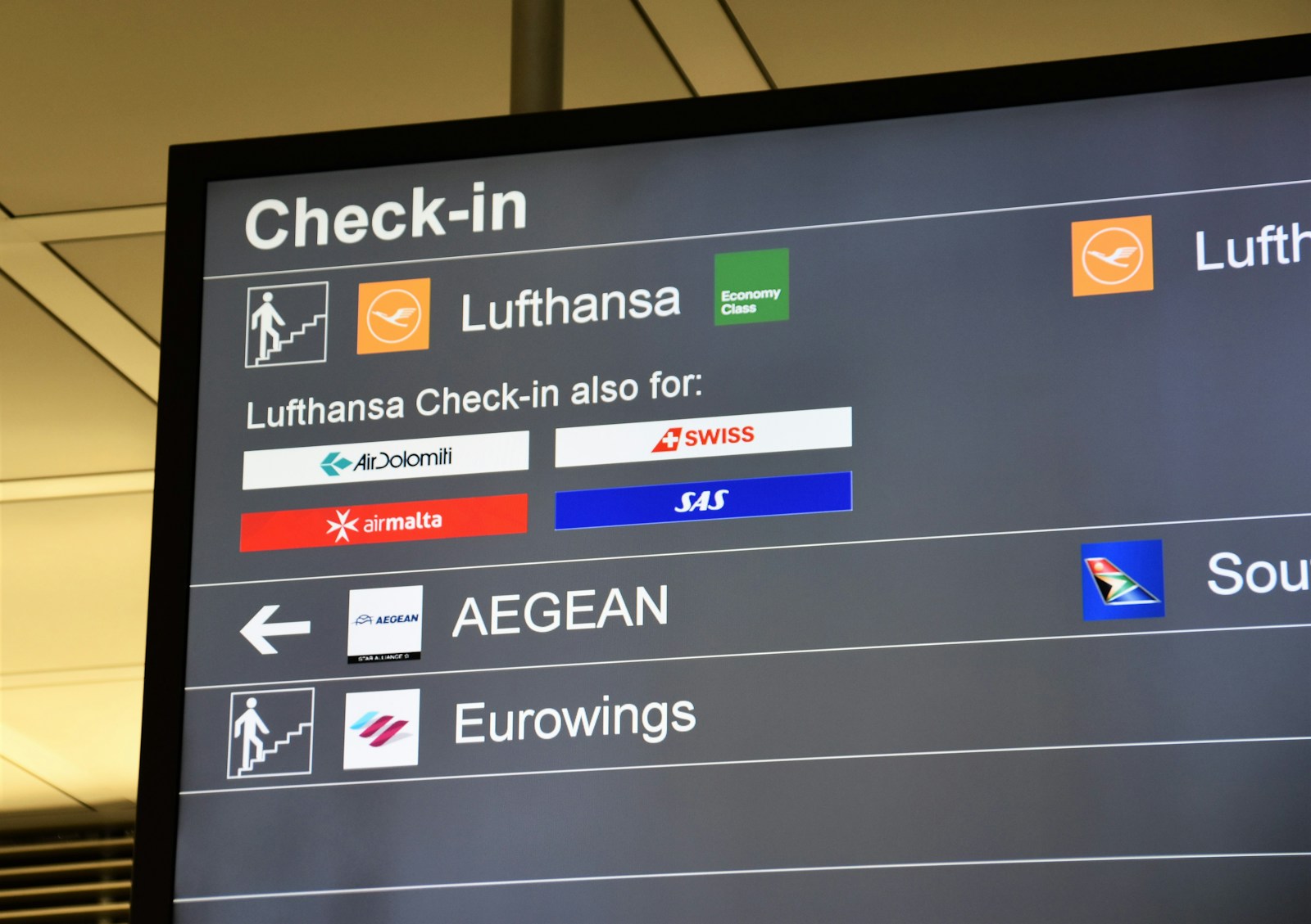 Painel de check-in de aeroporto mostrando logos de várias companhias aéreas parceiras, como Lufthansa, Swiss e Aegean