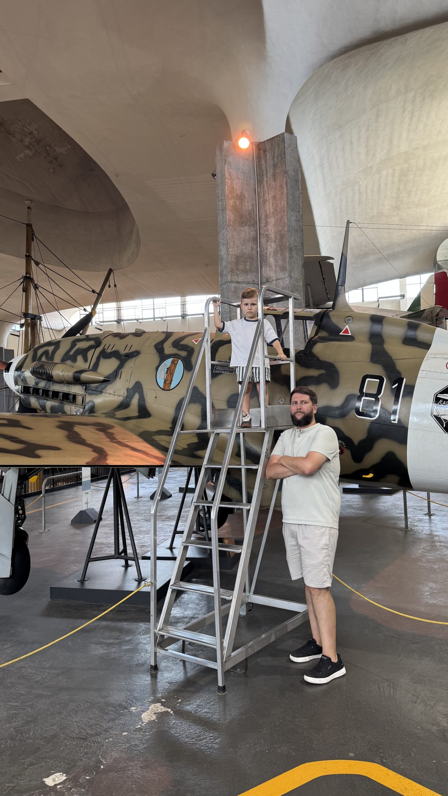 Pai e filho em frente a um avião de caça camuflado exposto no hangar de aviação do museu