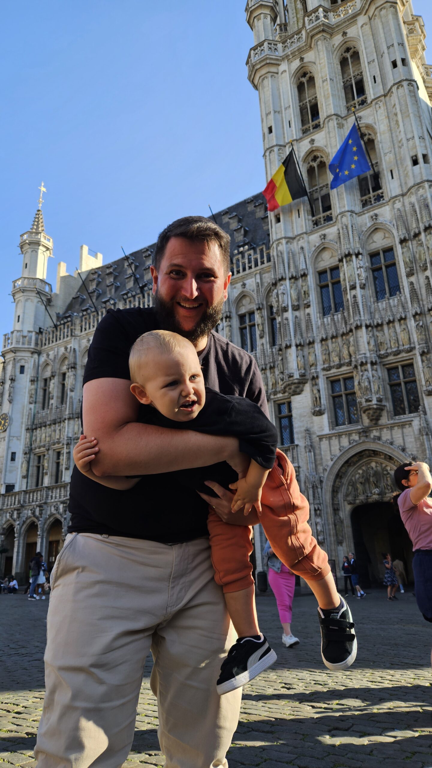 Pai segurando o bebe no colo com a fachada historica da Grand Place de Bruxelas ao fundo