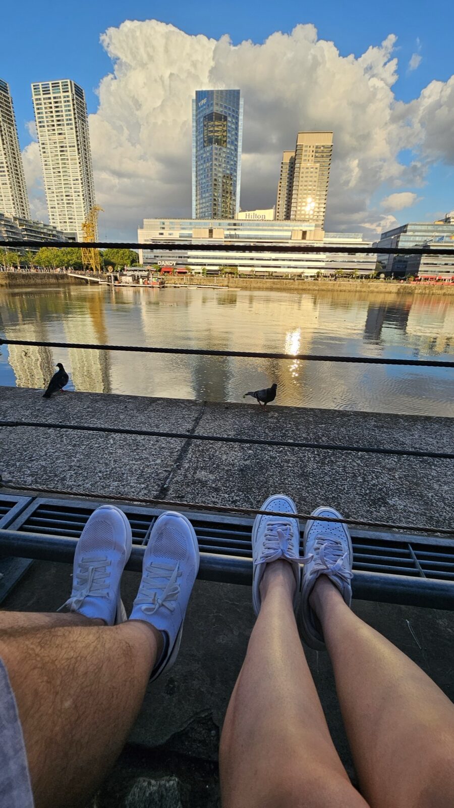 Pernas de duas pessoas sentadas na doca do Puerto Madero, com pombos na borda e o skyline ao fundo