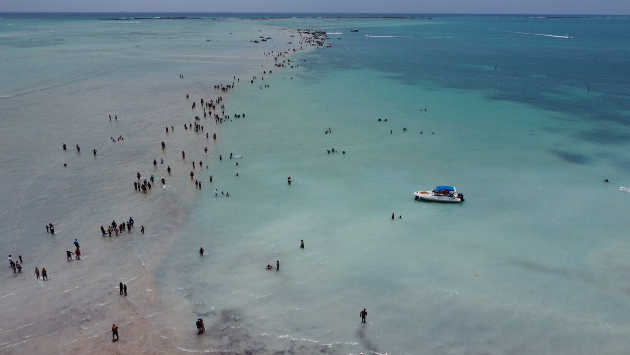 Vista aérea de drone do Caminho de Moisés em Barra Grande, Maragogi, com faixa de areia exposta na maré baixa e lancha ancorada ao lado.
