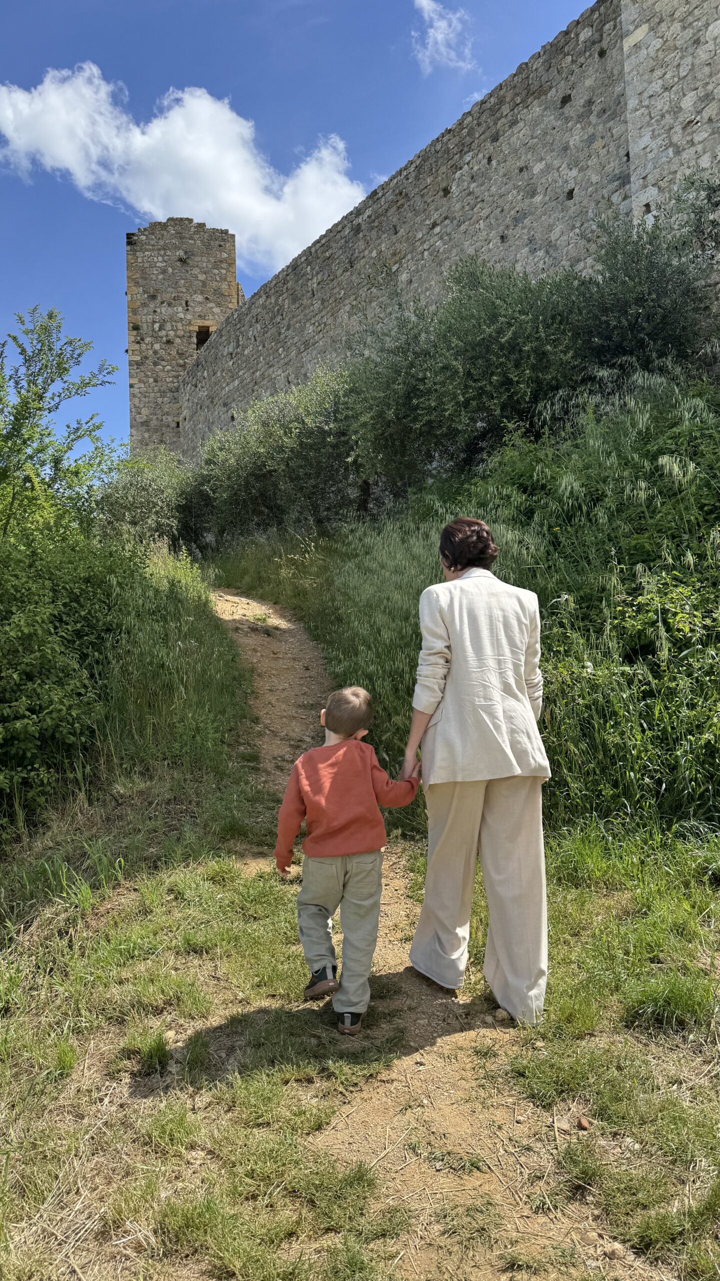 Caminho de terra ao lado externo das muralhas de Monteriggioni, com uma torre em ruínas ao fundo sob céu azul
