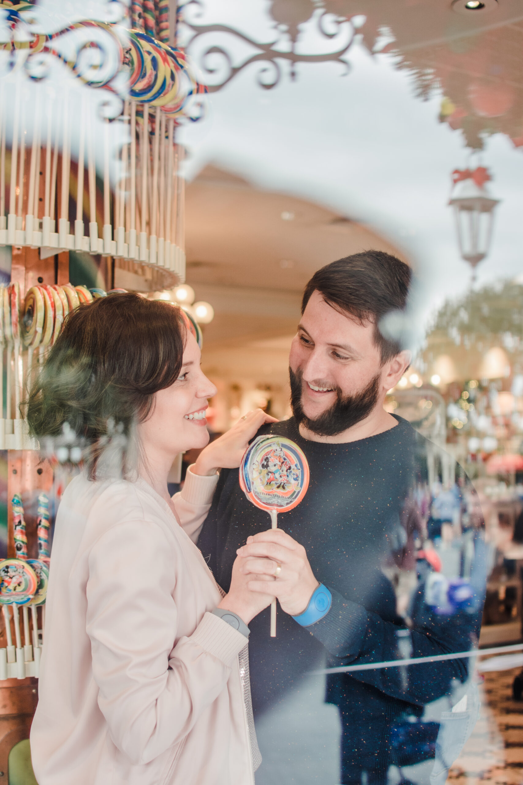 Casal sorrindo segurando pirulito da Disney refletido na vitrine da doceria da Main Street
