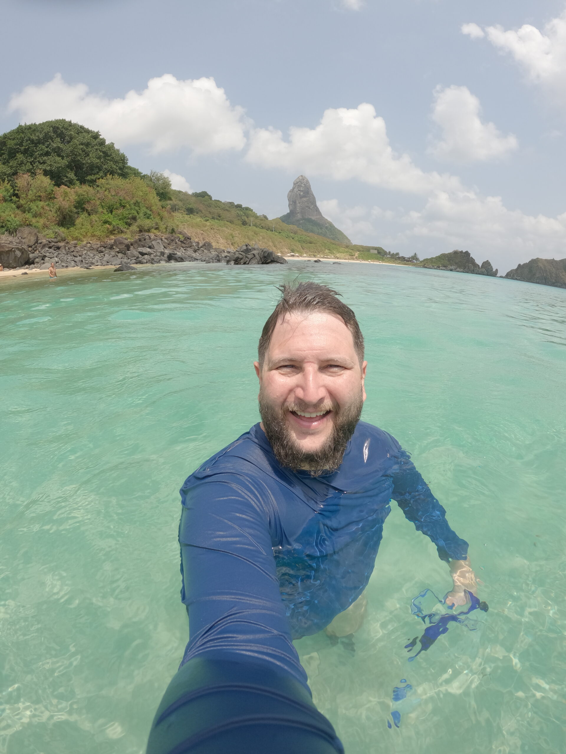 Homem sorrindo na água em Fernando de Noronha com o Morro do Pico ao fundo