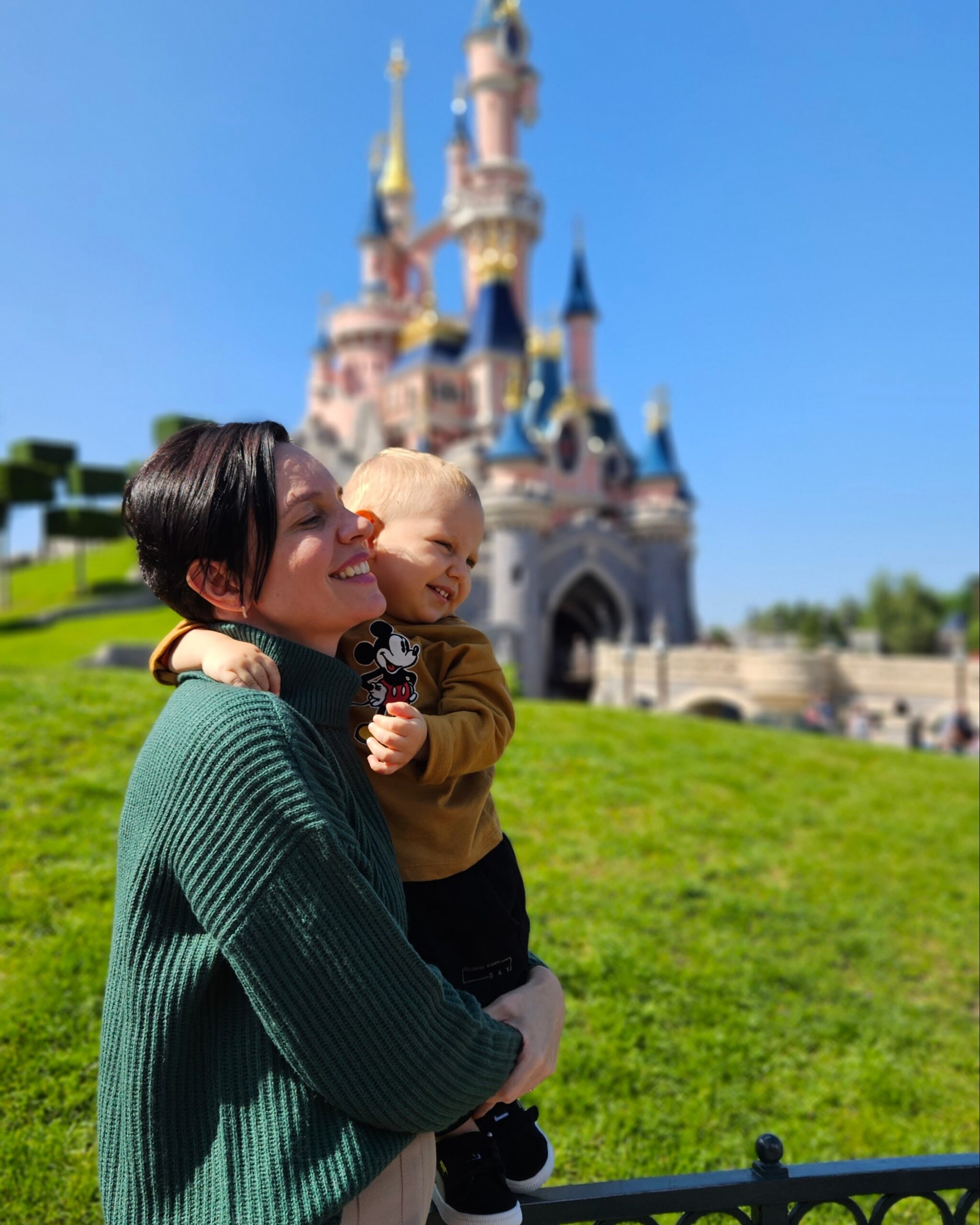 Mae abracada com o bebe sorrindo em frente ao castelo da Disneyland Paris