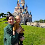 Mae segurando o bebe vestindo camiseta do Mickey em frente ao castelo da Bela Adormecida na Disneyland Paris