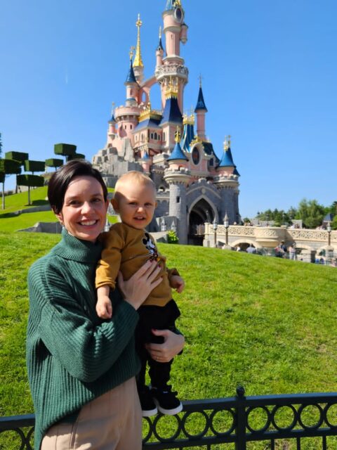 Mae segurando o bebe vestindo camiseta do Mickey em frente ao castelo da Bela Adormecida na Disneyland Paris
