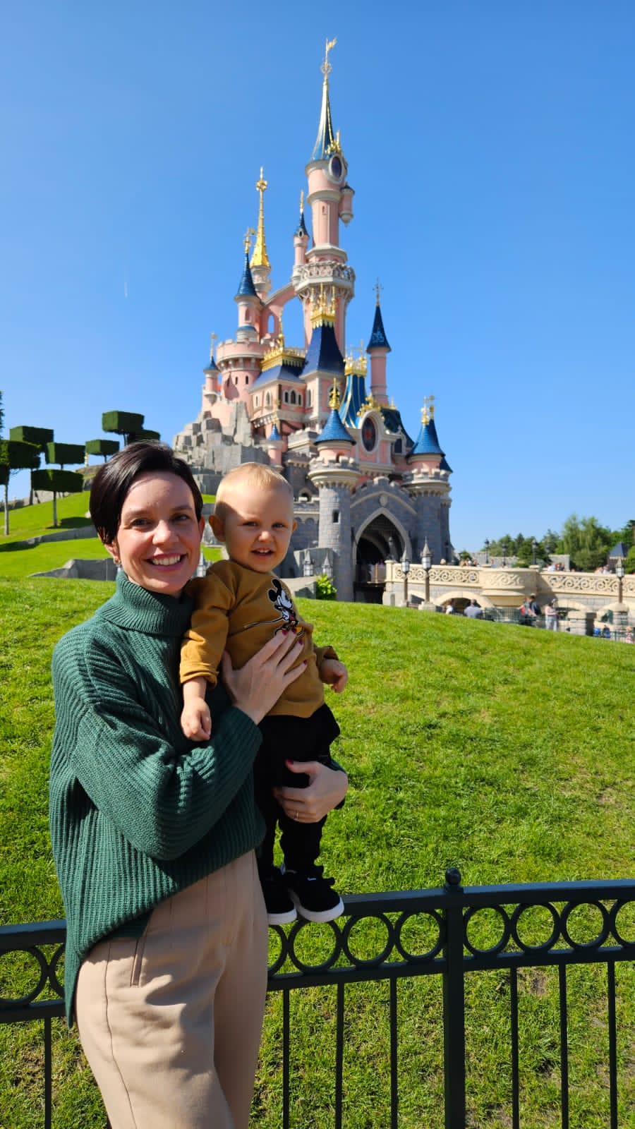 Mae segurando o bebe vestindo camiseta do Mickey em frente ao castelo da Bela Adormecida na Disneyland Paris