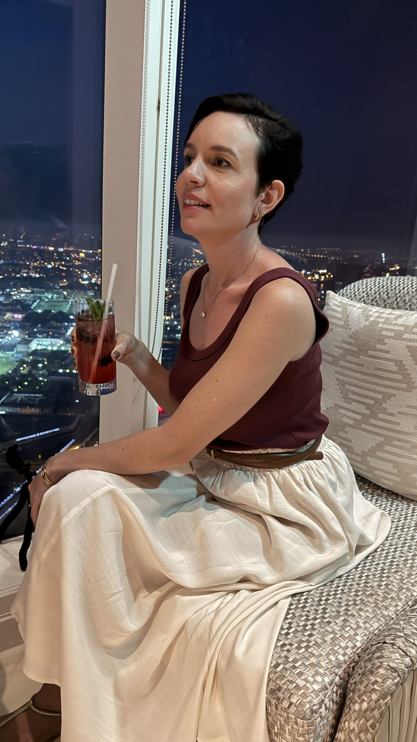 Carol com um drinque à noite, com vista da skyline de Dubai iluminada ao fundo