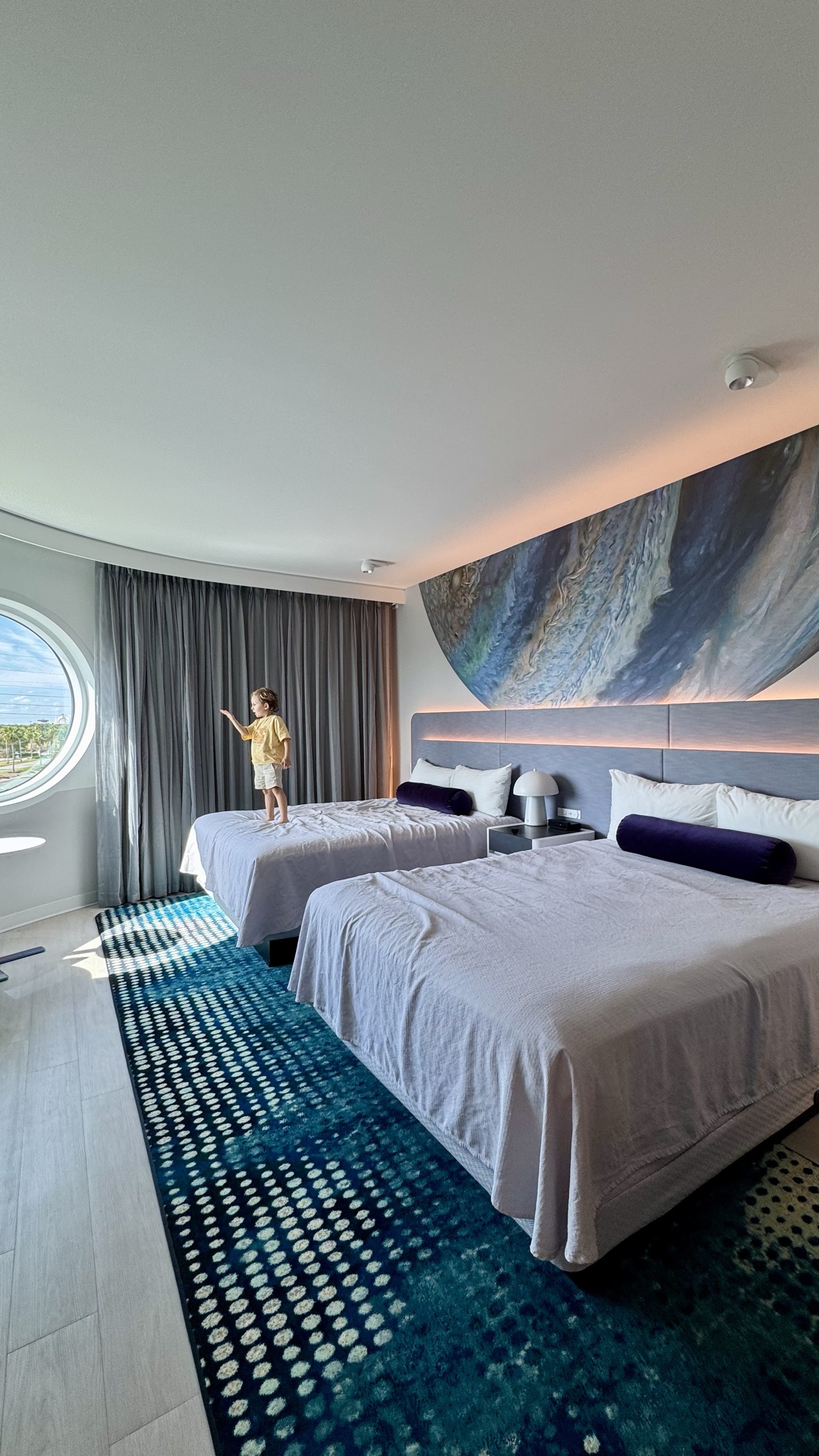 Crianca em pe na cama do quarto do hotel Terra Luna, olhando pela janela redonda na manha da visita ao Epic Universe