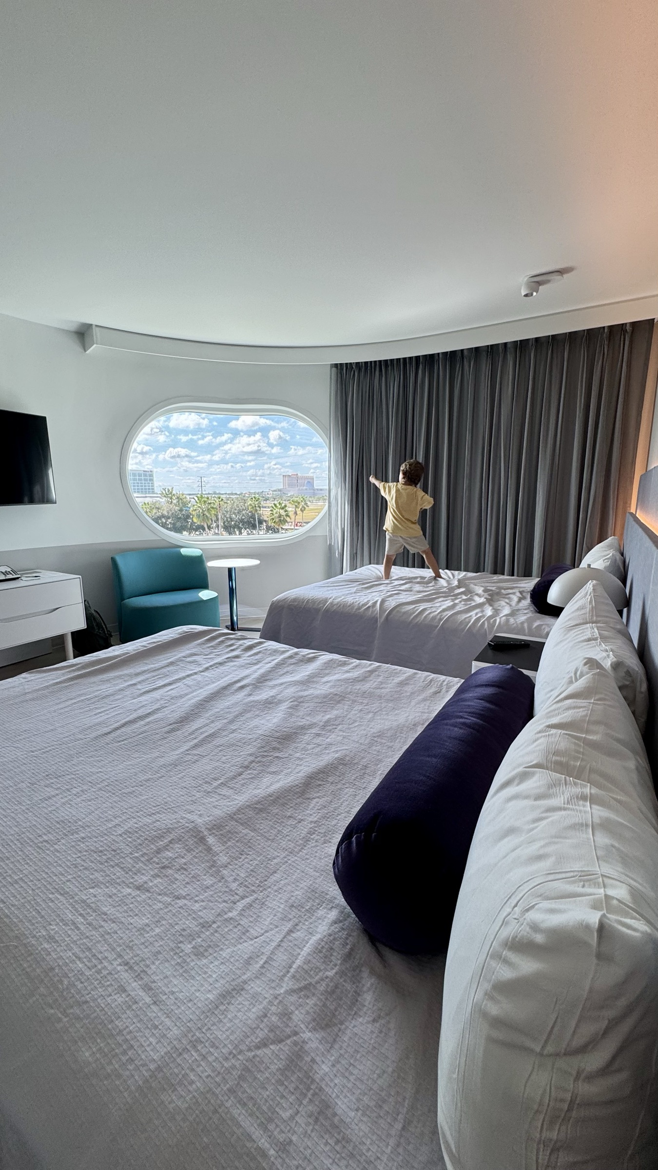 Samuel em pe na cama do quarto do hotel Terra Luna, olhando pela janela na manha da visita ao Epic Universe