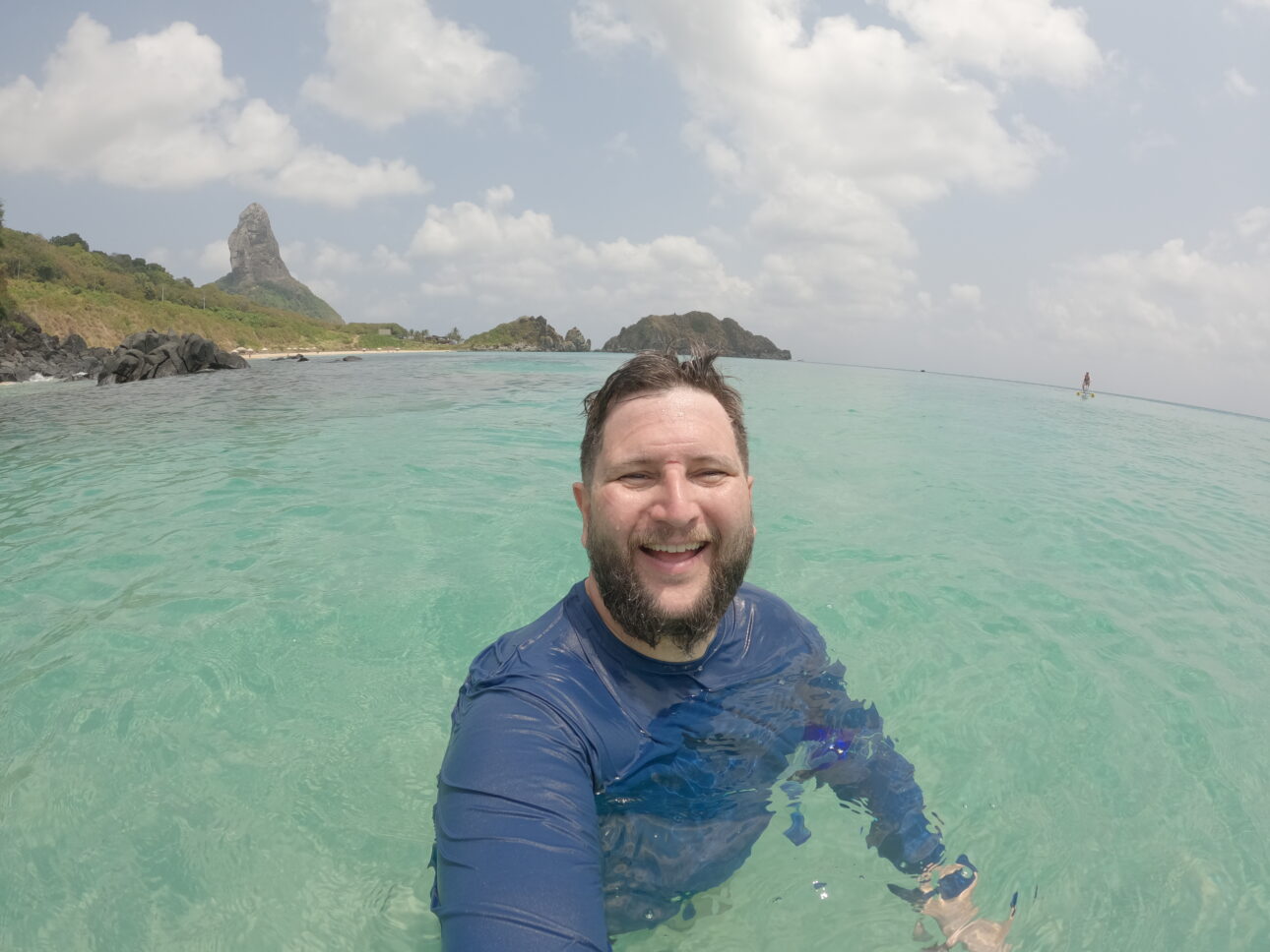 Homem sorrindo dentro do mar turquesa de Fernando de Noronha, com o Morro do Pico ao fundo