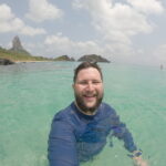 Homem sorrindo dentro do mar turquesa de Fernando de Noronha, com o Morro do Pico ao fundo