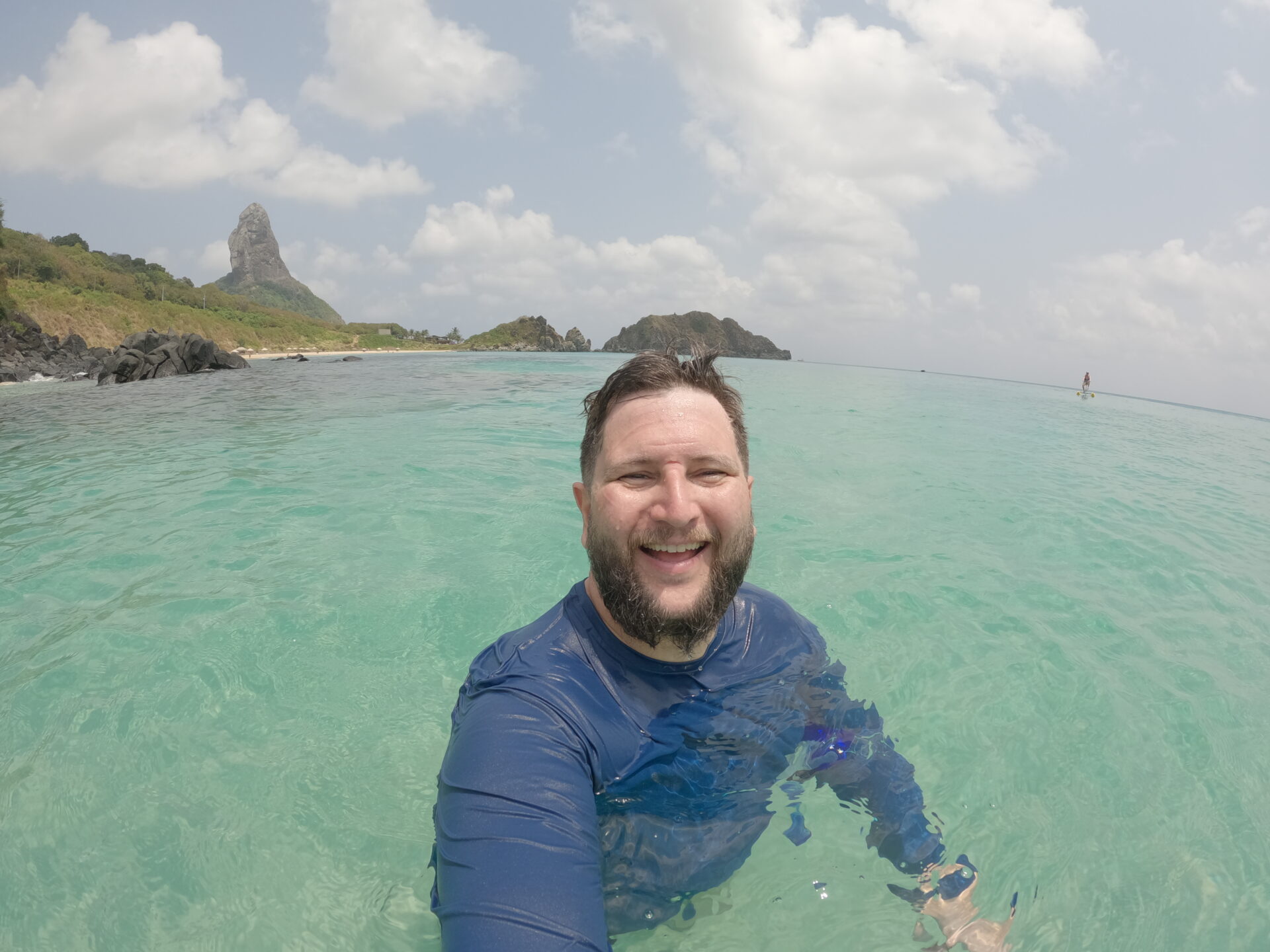 Homem sorrindo dentro do mar turquesa de Fernando de Noronha, com o Morro do Pico ao fundo