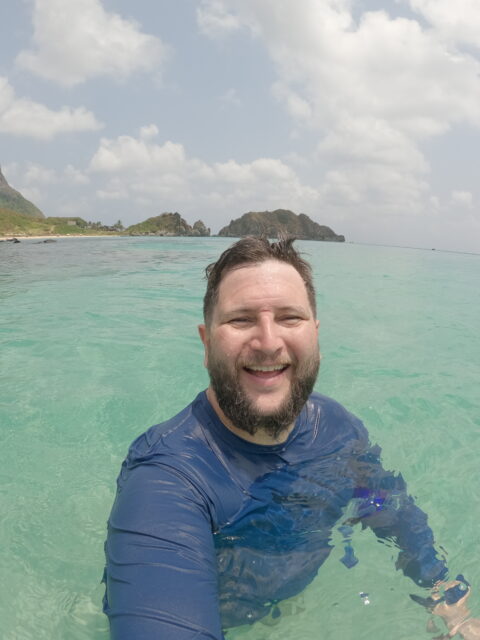 Homem sorrindo dentro do mar turquesa de Fernando de Noronha, com o Morro do Pico ao fundo