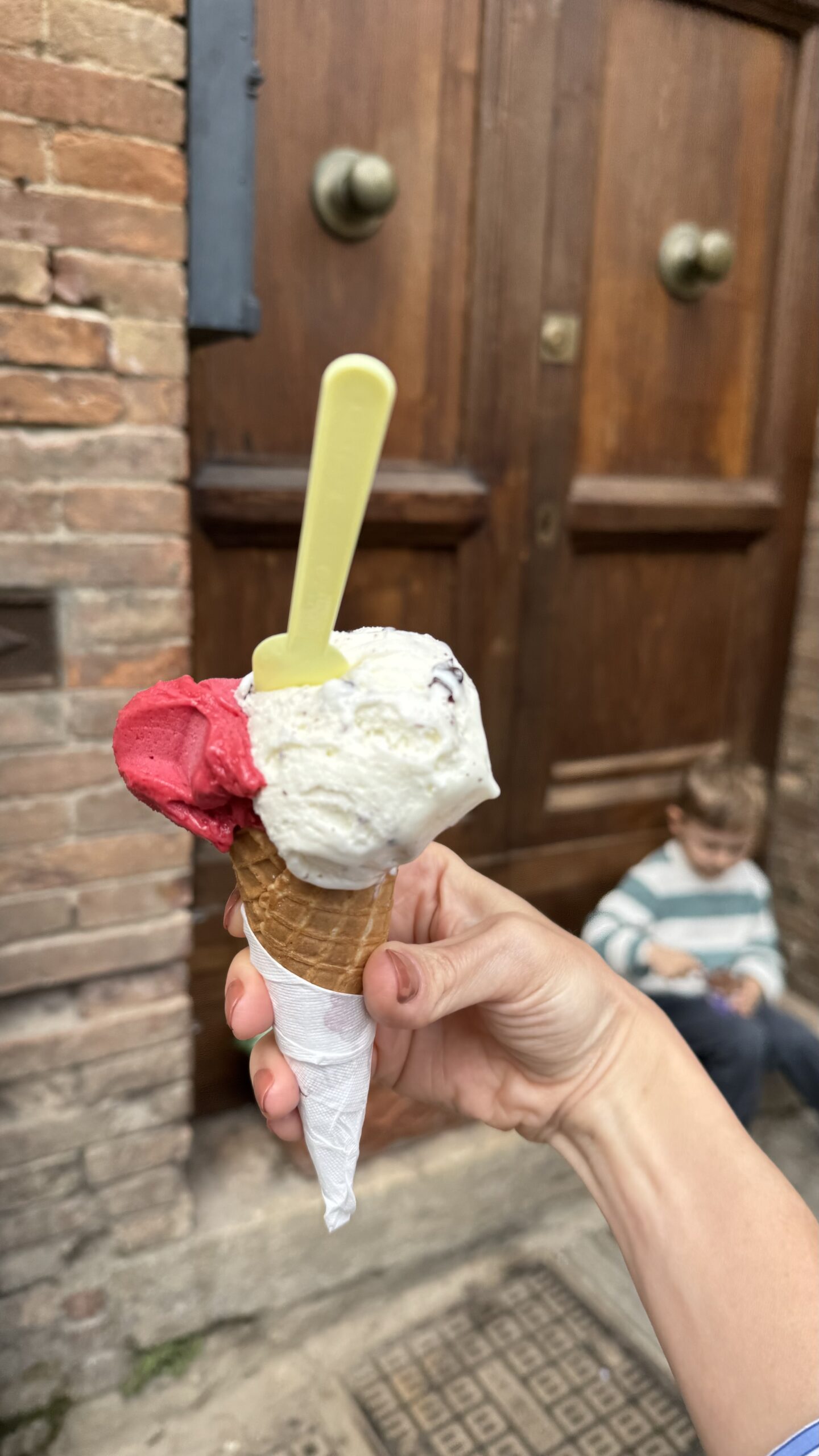 Casquinha de gelato com duas bolas, uma branca e uma vermelha, segurada na mão em frente a uma porta de madeira