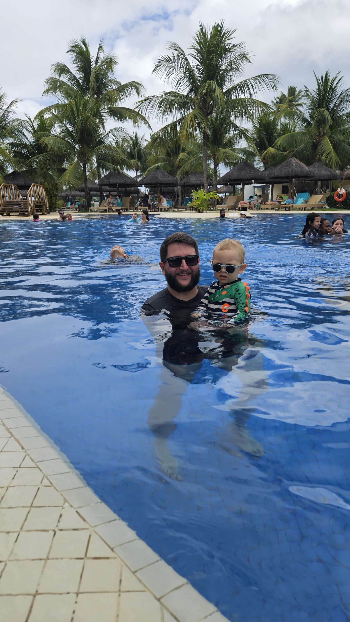 Rudi com o Samuel de óculos escuros na piscina do Iberostar Praia do Forte cercada de coqueiros