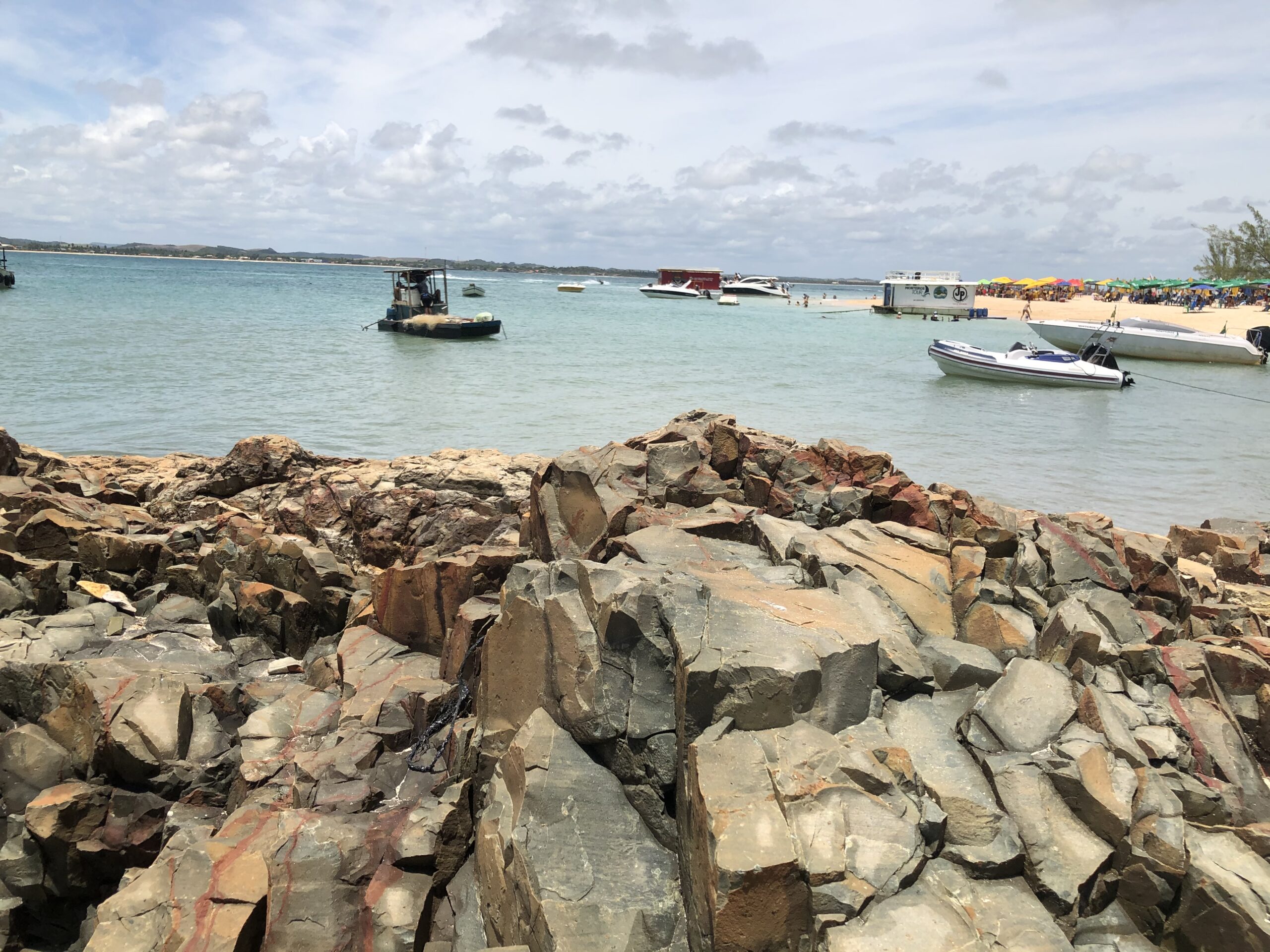 Lanchas ancoradas próximas à praia principal da Ilha de Santo Aleixo, vistas das pedras da costa, com guarda-sóis coloridos ao fundo.