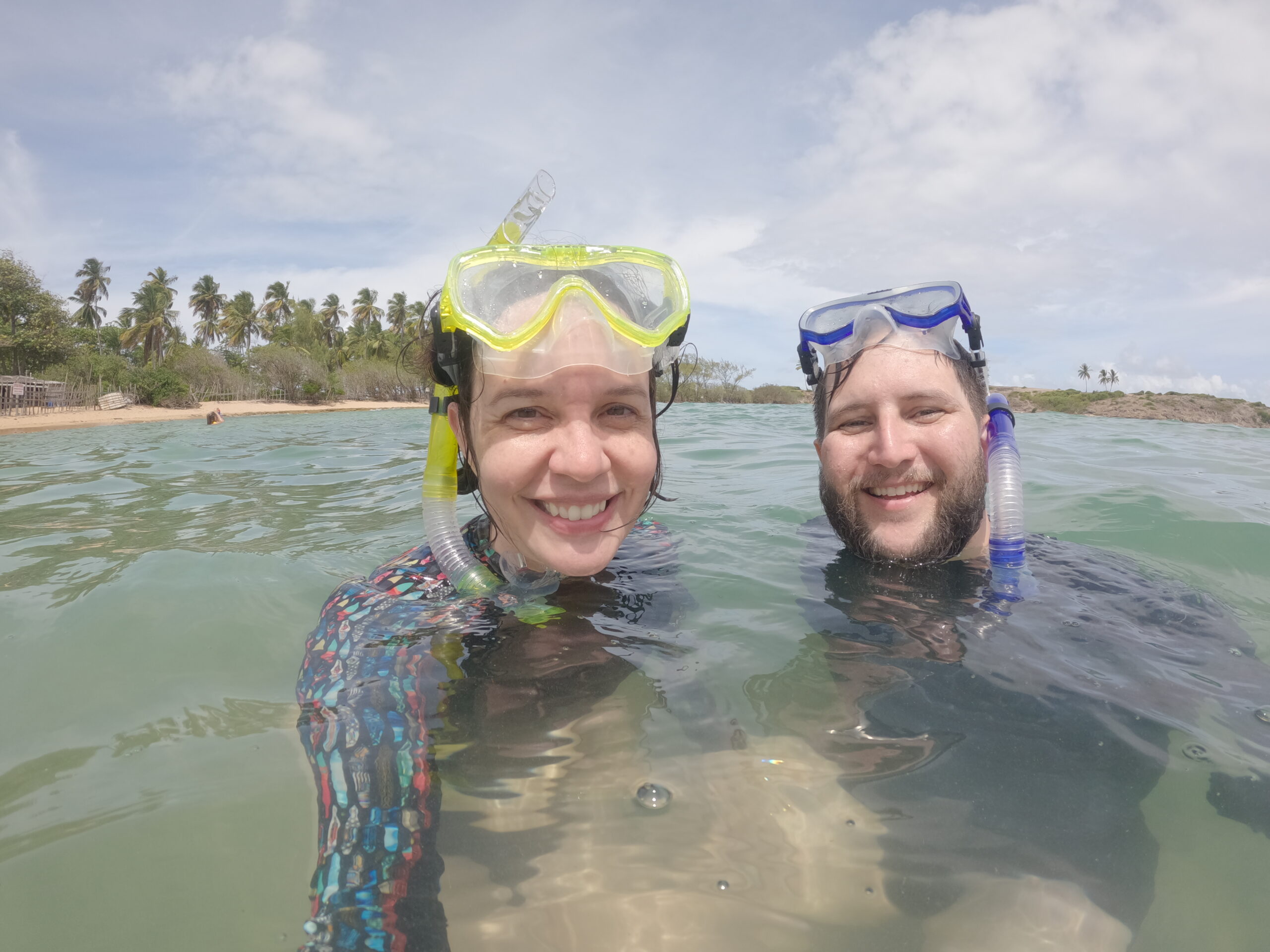 Casal com máscara e snorkel dentro da água na Praia da Ferradura, com a faixa de coqueiros da Ilha de Santo Aleixo ao fundo.