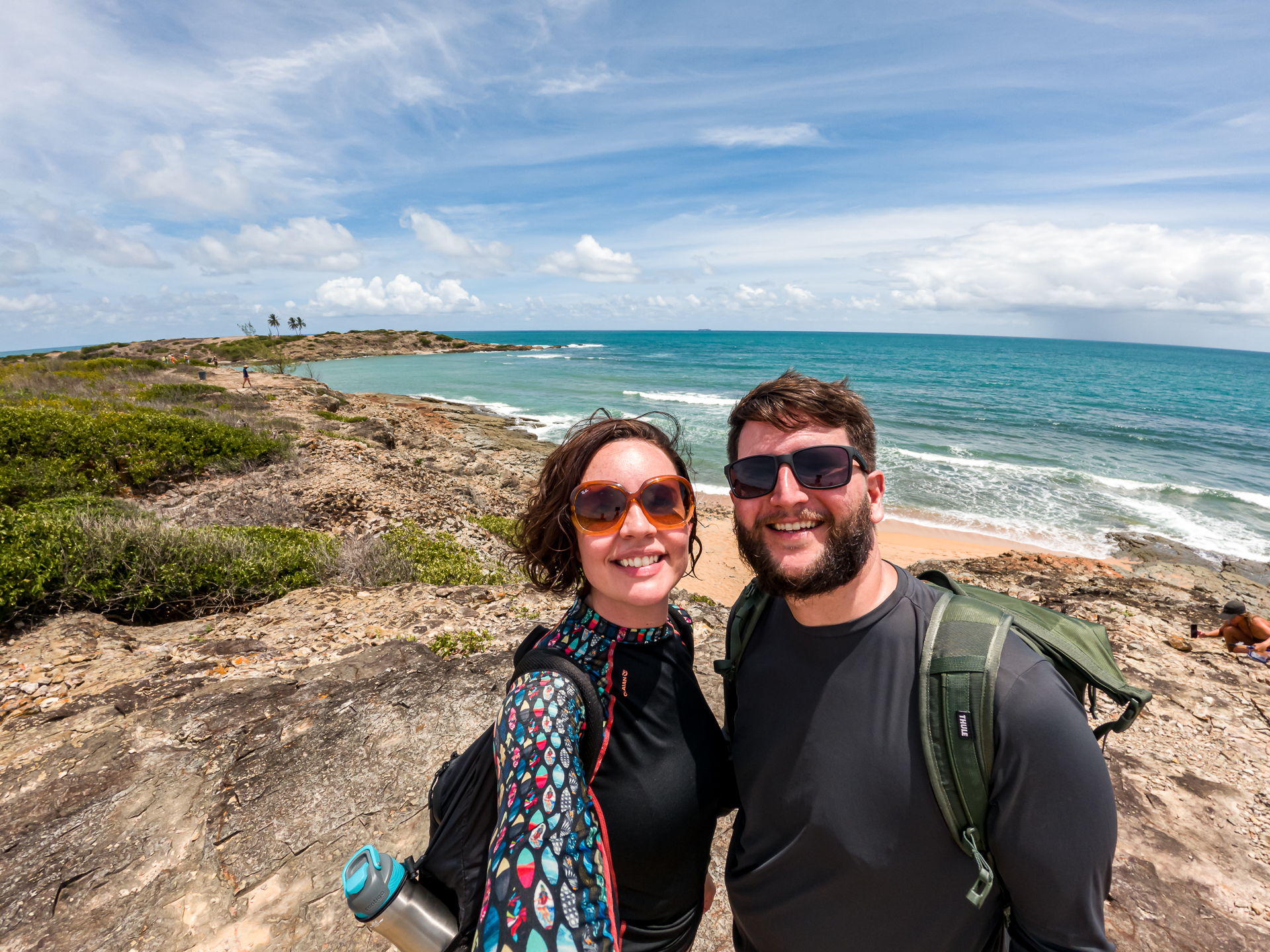 Casal com mochilas no ponto mais alto da trilha da Ilha de Santo Aleixo, com vista panorâmica da costa rochosa e do mar.