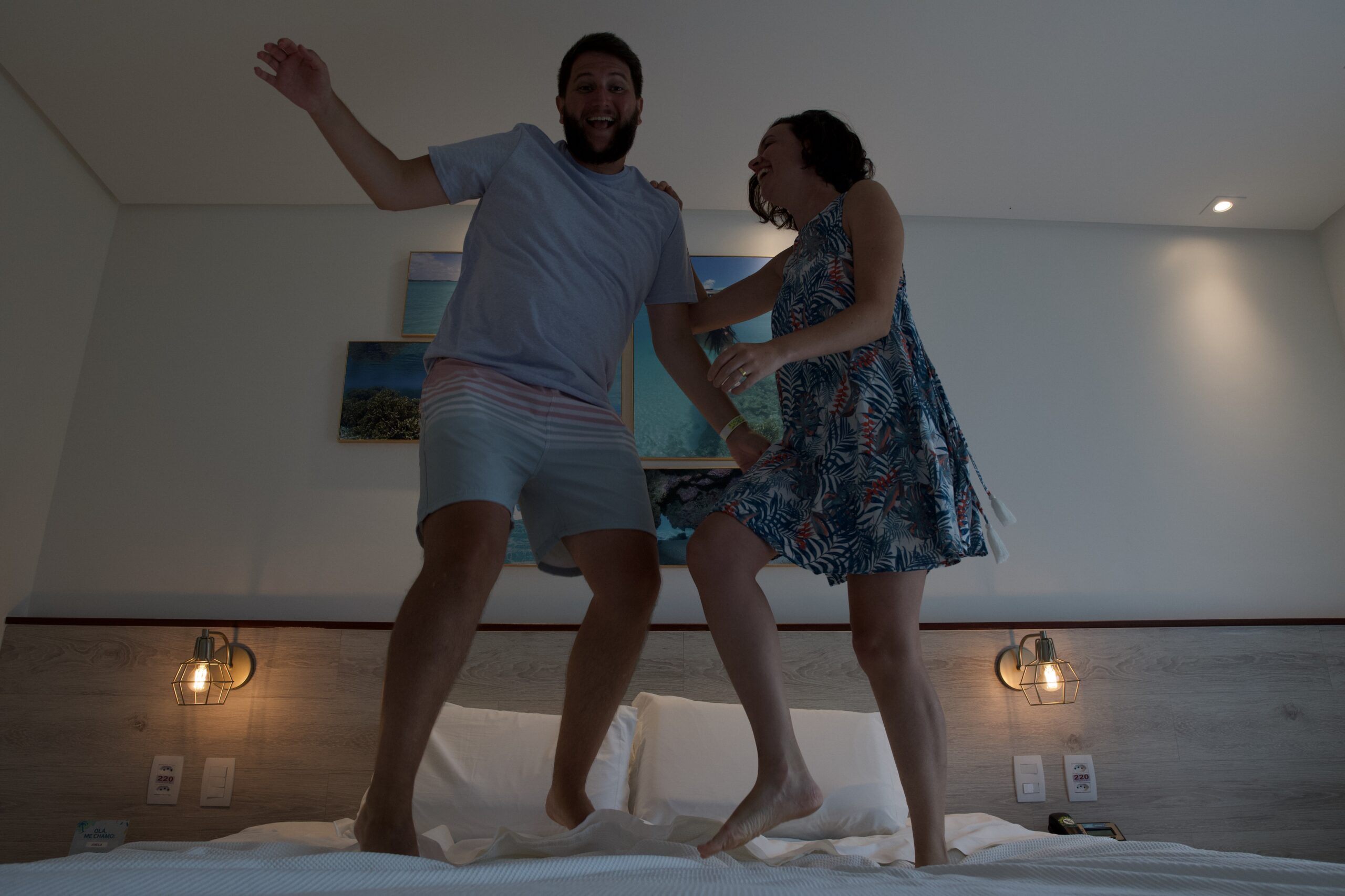 Casal pulando na cama do quarto Standard do Japaratinga Lounge Resort, com luminárias e quadros de coral na parede