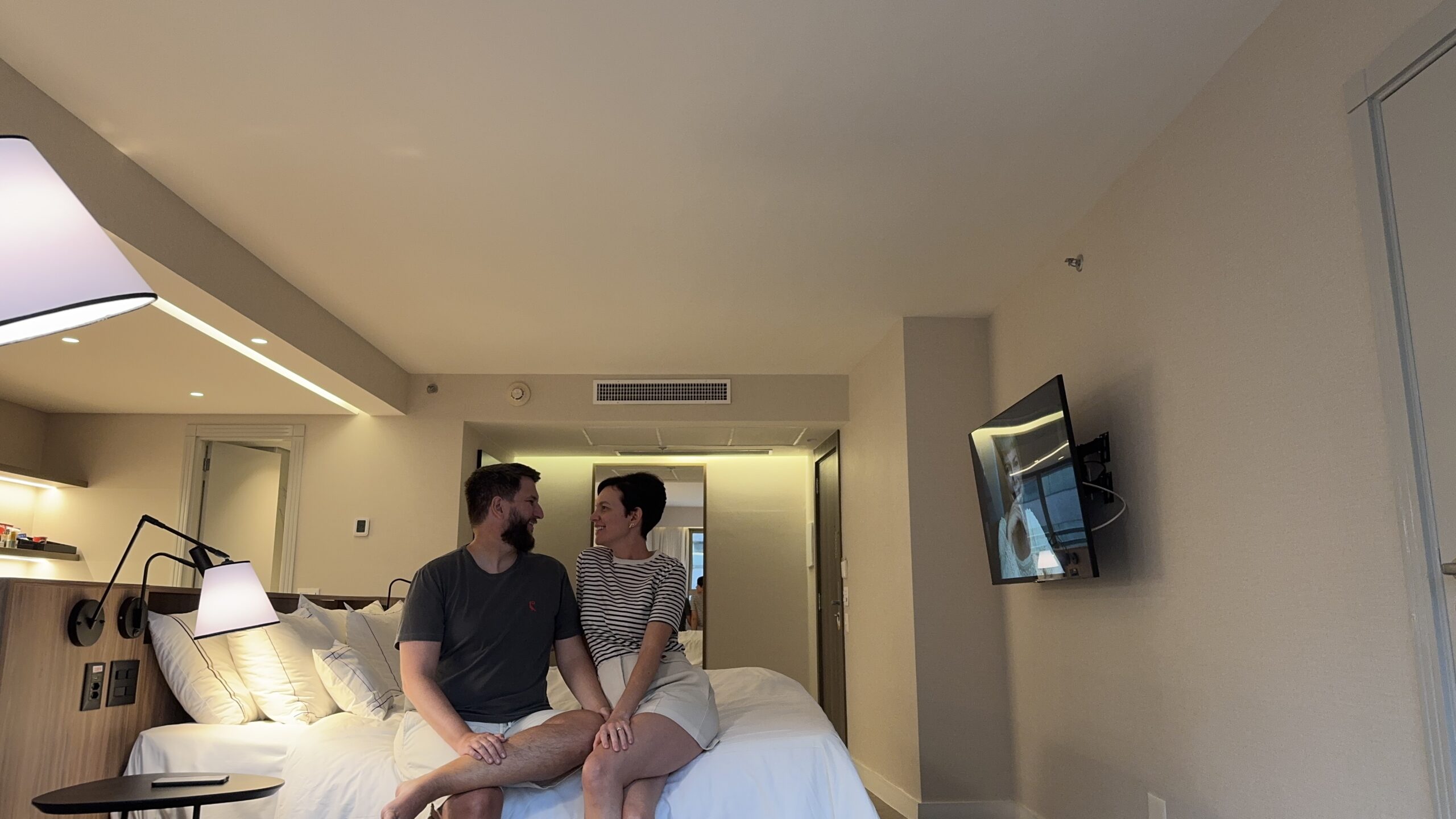 Casal sentado na cama do quarto The Level do Meliá Brasil 21 com a TV ao fundo