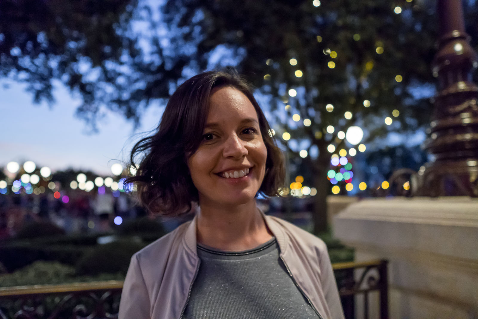 Mulher de jaqueta leve sorrindo a noite no Magic Kingdom com luzes desfocadas ao fundo
