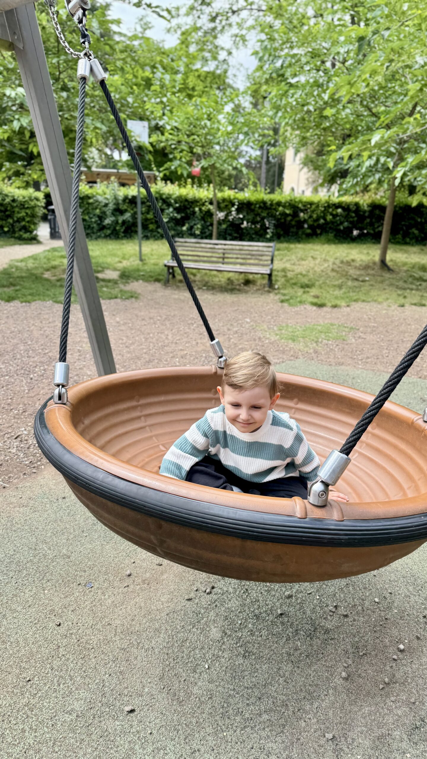 Criança brincando em um balanço no parquinho da Villa Borghese, cercado de árvores e um banco de praça.