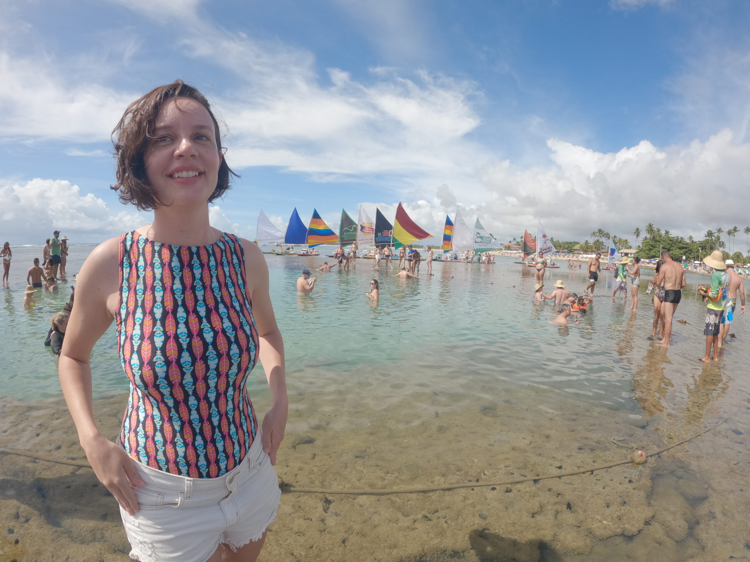 Mulher sorridente em pe dentro de uma piscina natural em Porto de Galinhas, com jangadas coloridas ancoradas ao fundo
