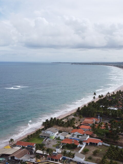 Vista aerea da praia de Porto de Galinhas, com casas de telhado alaranjado na orla e o mar em tons de azul e verde