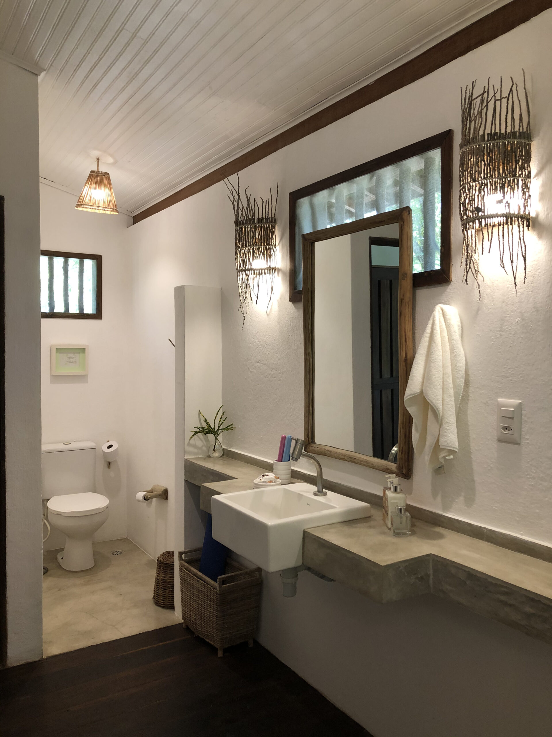 Banheiro do bangalo com bancada de cimento queimado, espelho de madeira, toalhas dobradas e vaso sanitario