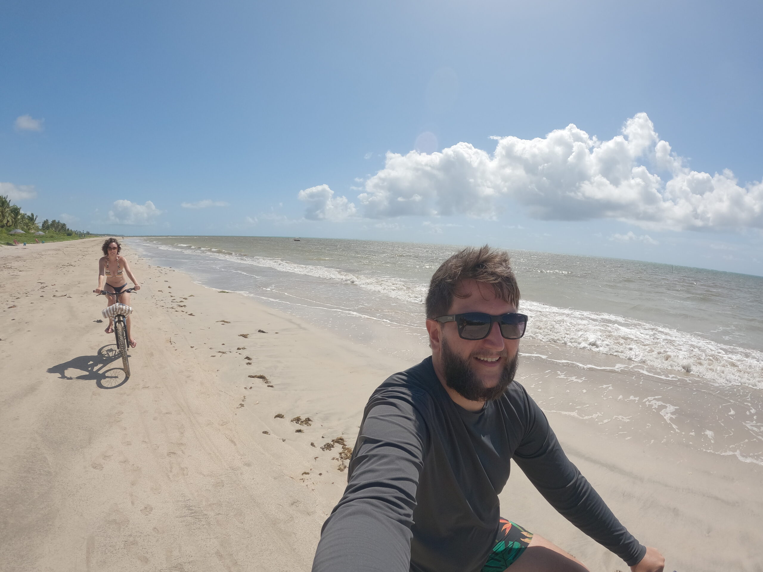 Casal andando de bicicleta na faixa de areia em frente a Praia do Toque, com o mar ao fundo