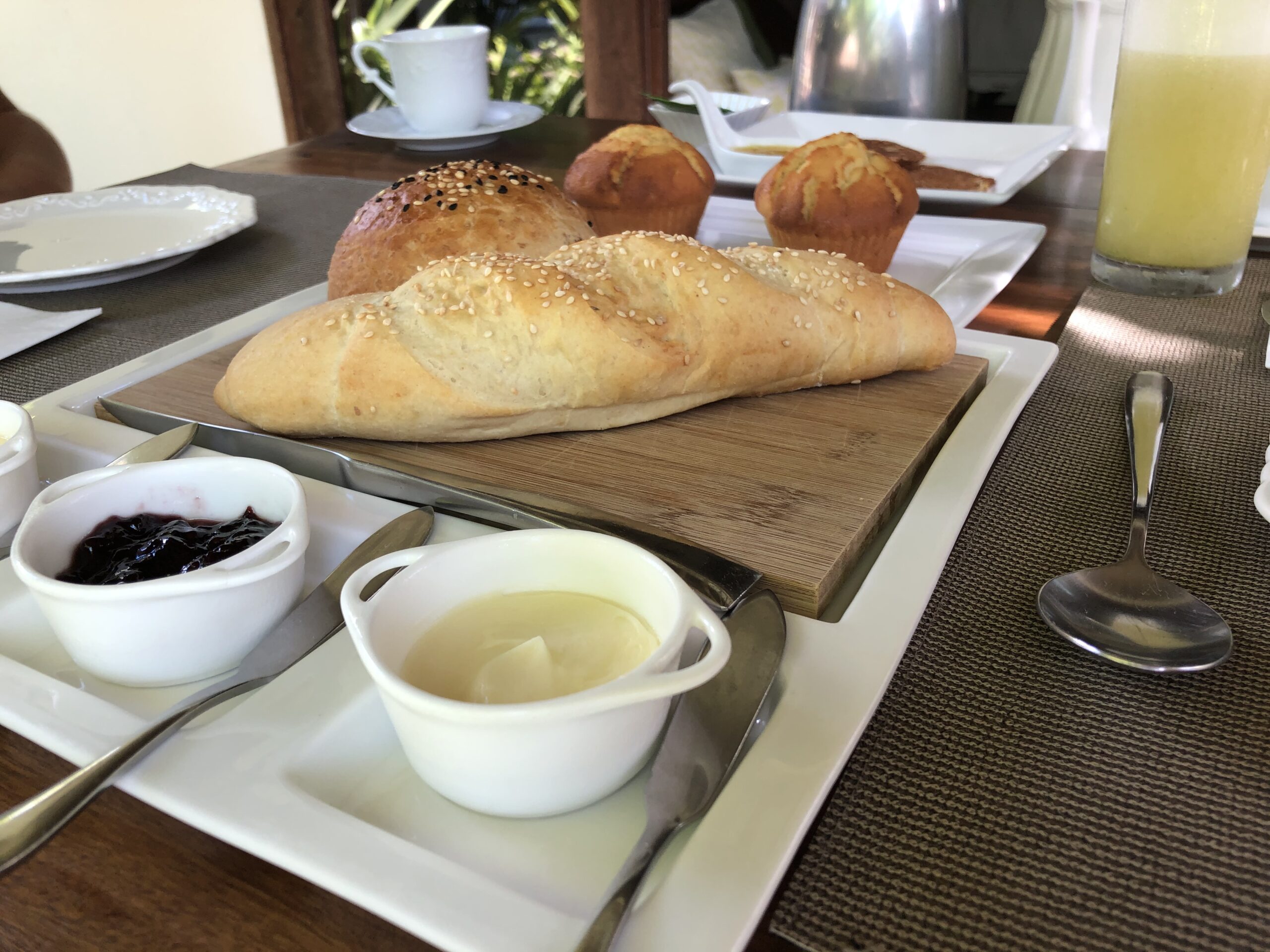 Mesa do cafe da manha da pousada com paes quentinhos, muffins, geleia, manteiga e suco servidos em etapas