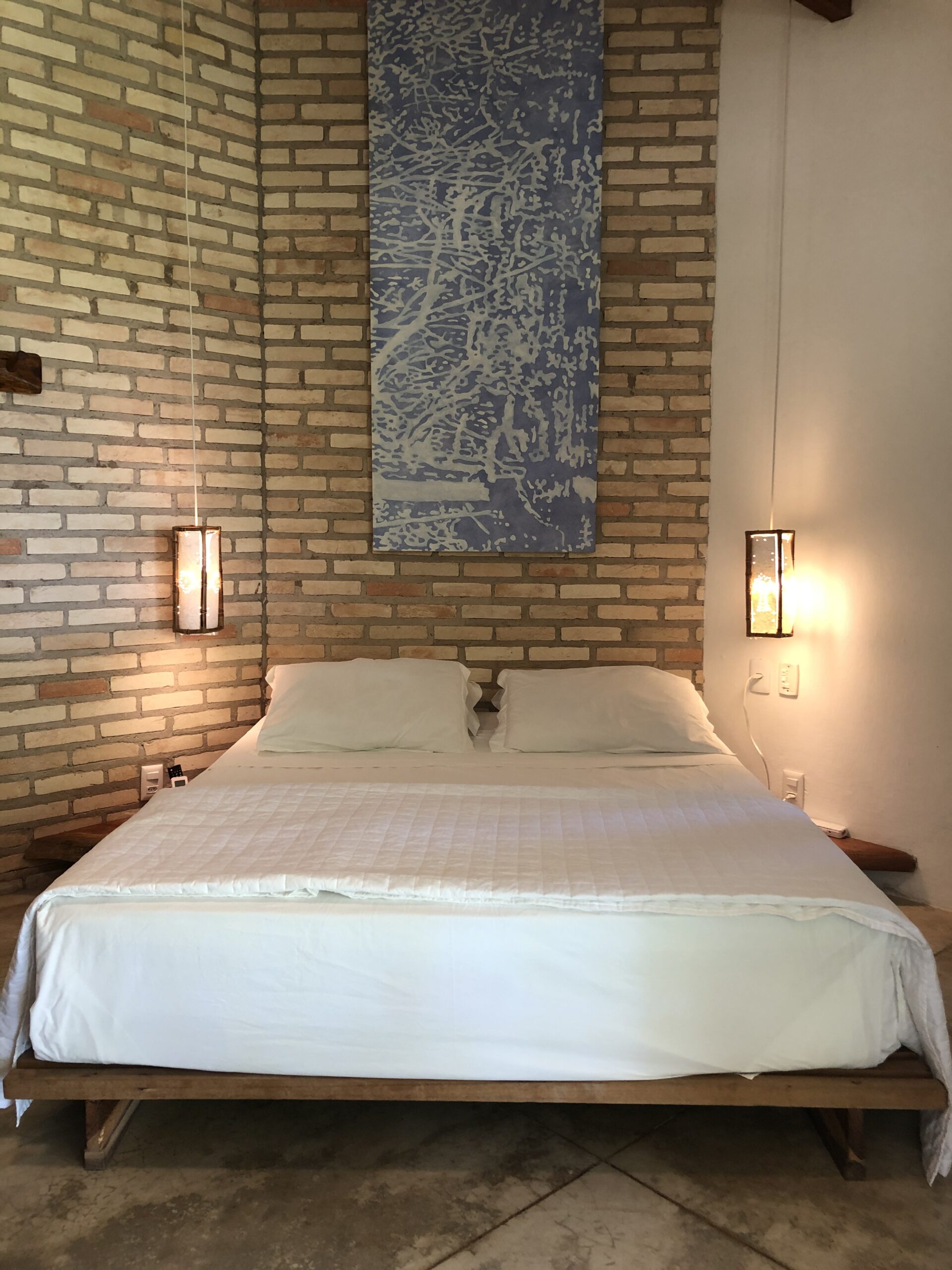 Quarto do bangalo Amendoeira com cama baixa sobre estrado, parede em tijolo aparente e quadro azul acima da cabeceira