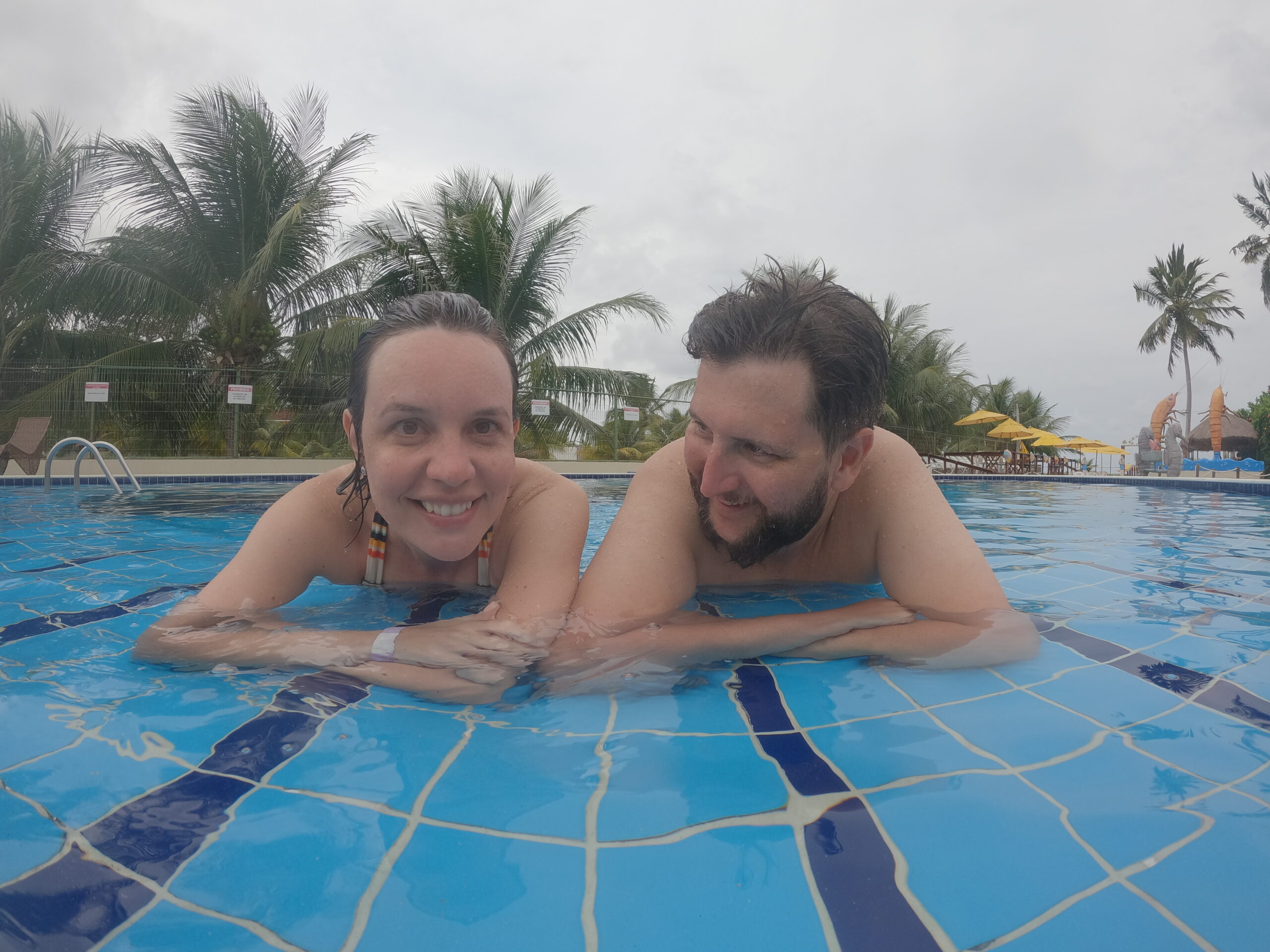 Casal na piscina da Pousada Polymar com coqueiros e guarda-sois amarelos ao fundo