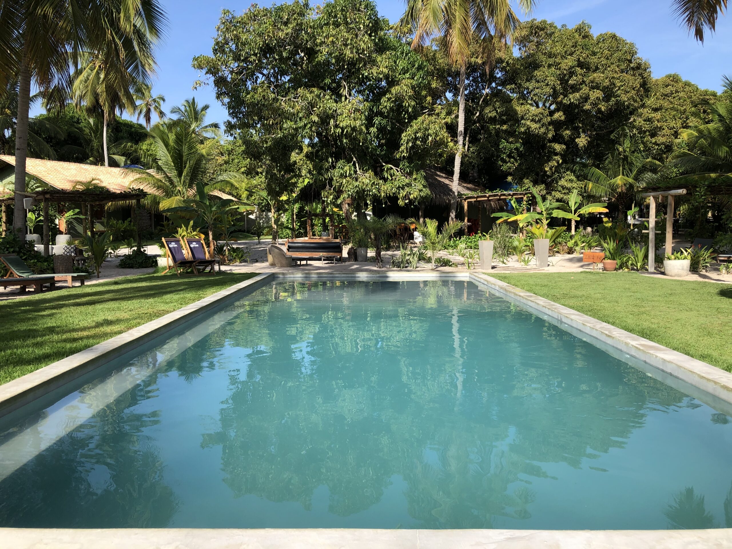 Piscina principal de cimento queimado da Pousada Quadrado de Charme cercada de gramado e coqueiros na area externa