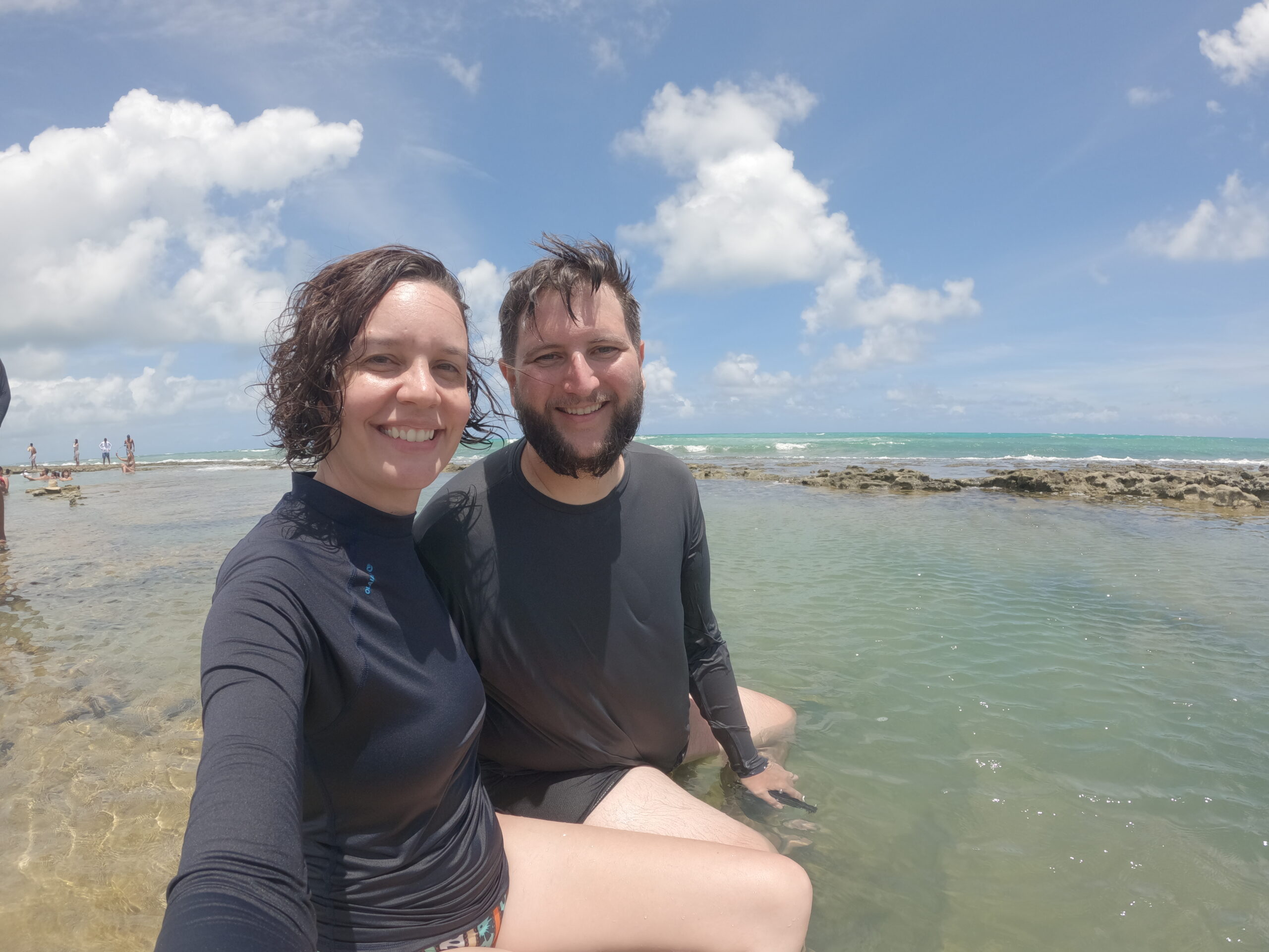 Casal sentado na água transparente e rasa da Praia dos Carneiros com o mar ao fundo
