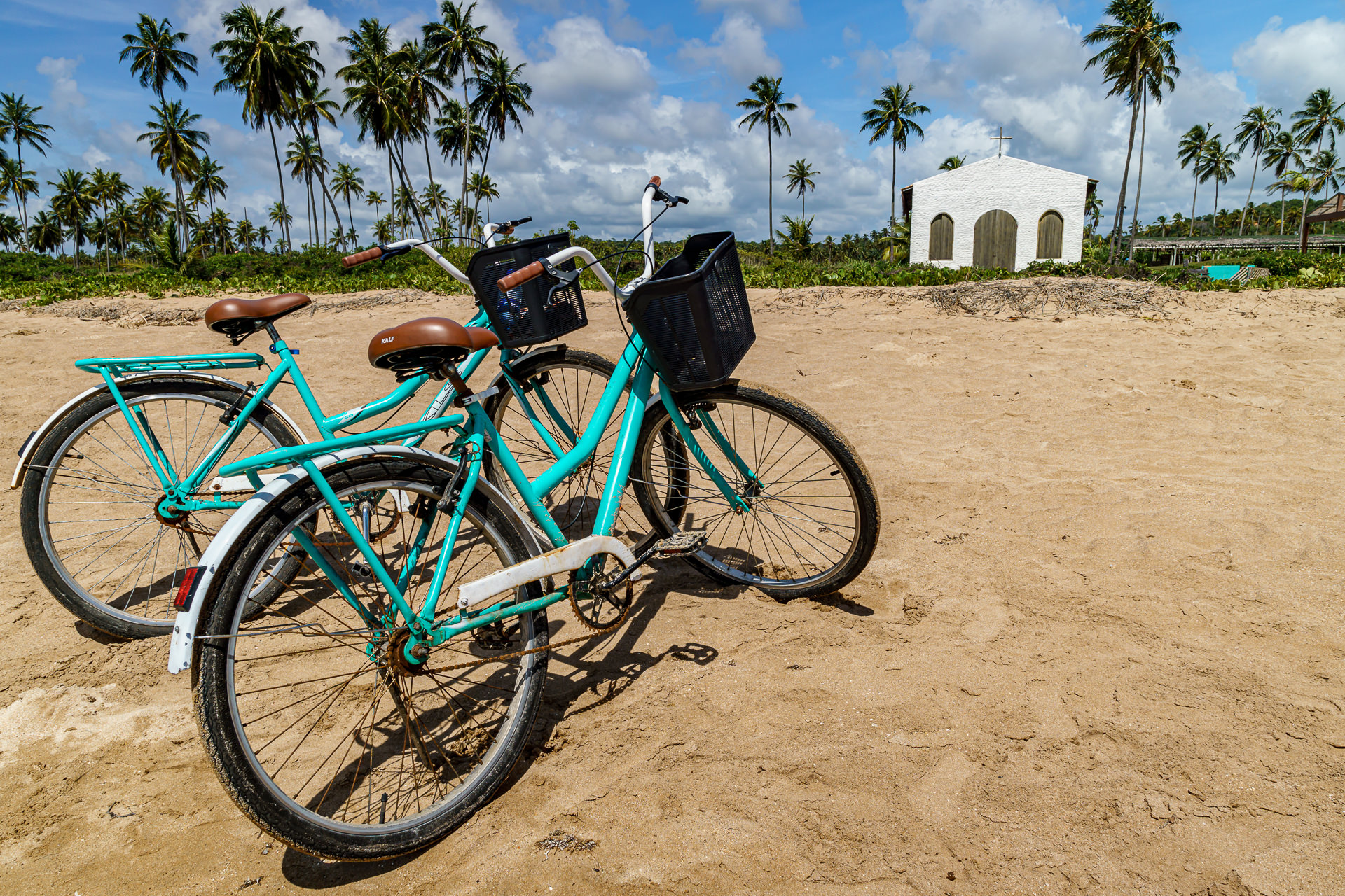 Duas bicicletas turquesa emprestadas pela pousada estacionadas na areia da Praia do Marceneiro com a Capela dos Milagres ao fundo
