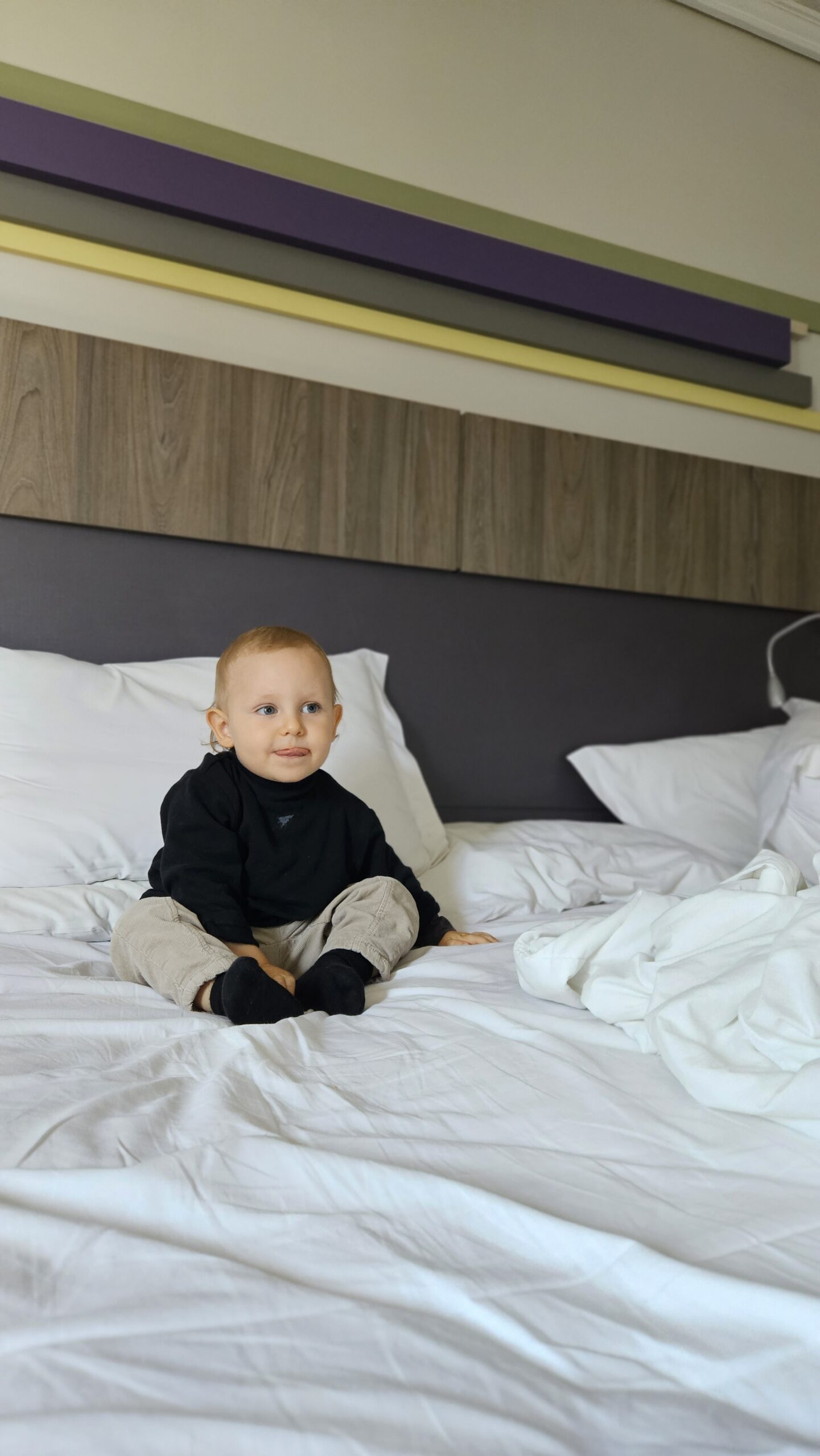 Sam em pe na cama do quarto do Grand Mercure Curitiba, com cabeceira listrada em roxo, verde e amarelo