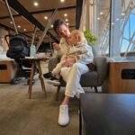 Carol com Samuel no colo sentados na Sala VIP BRB do Aeroporto de Brasília, com luz da manhã entrando pelo vidro