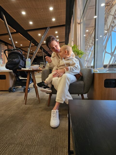 Carol com Samuel no colo sentados na Sala VIP BRB do Aeroporto de Brasília, com luz da manhã entrando pelo vidro