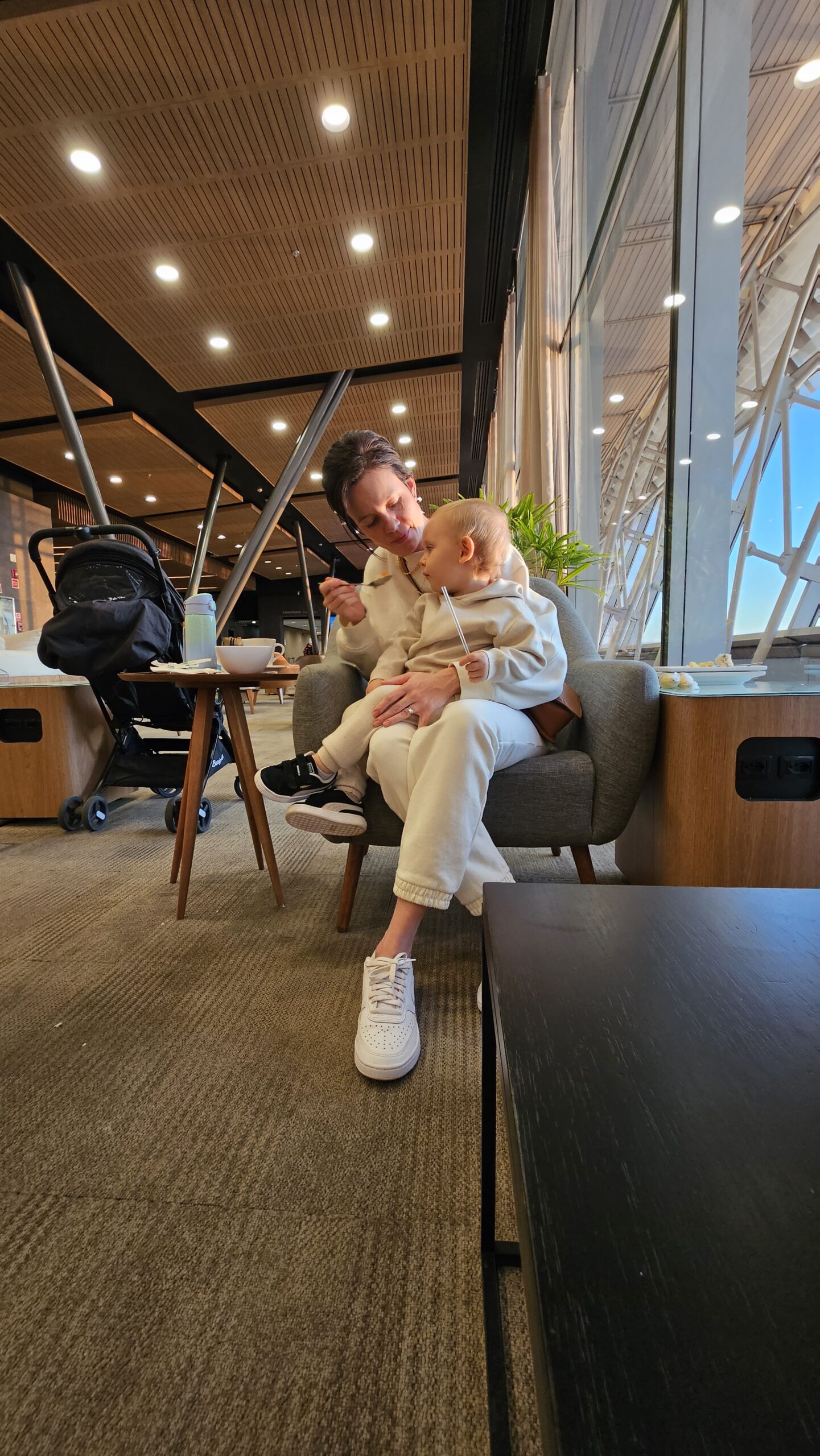 Carol com Samuel no colo sentados na Sala VIP BRB do Aeroporto de Brasília, com luz da manhã entrando pelo vidro