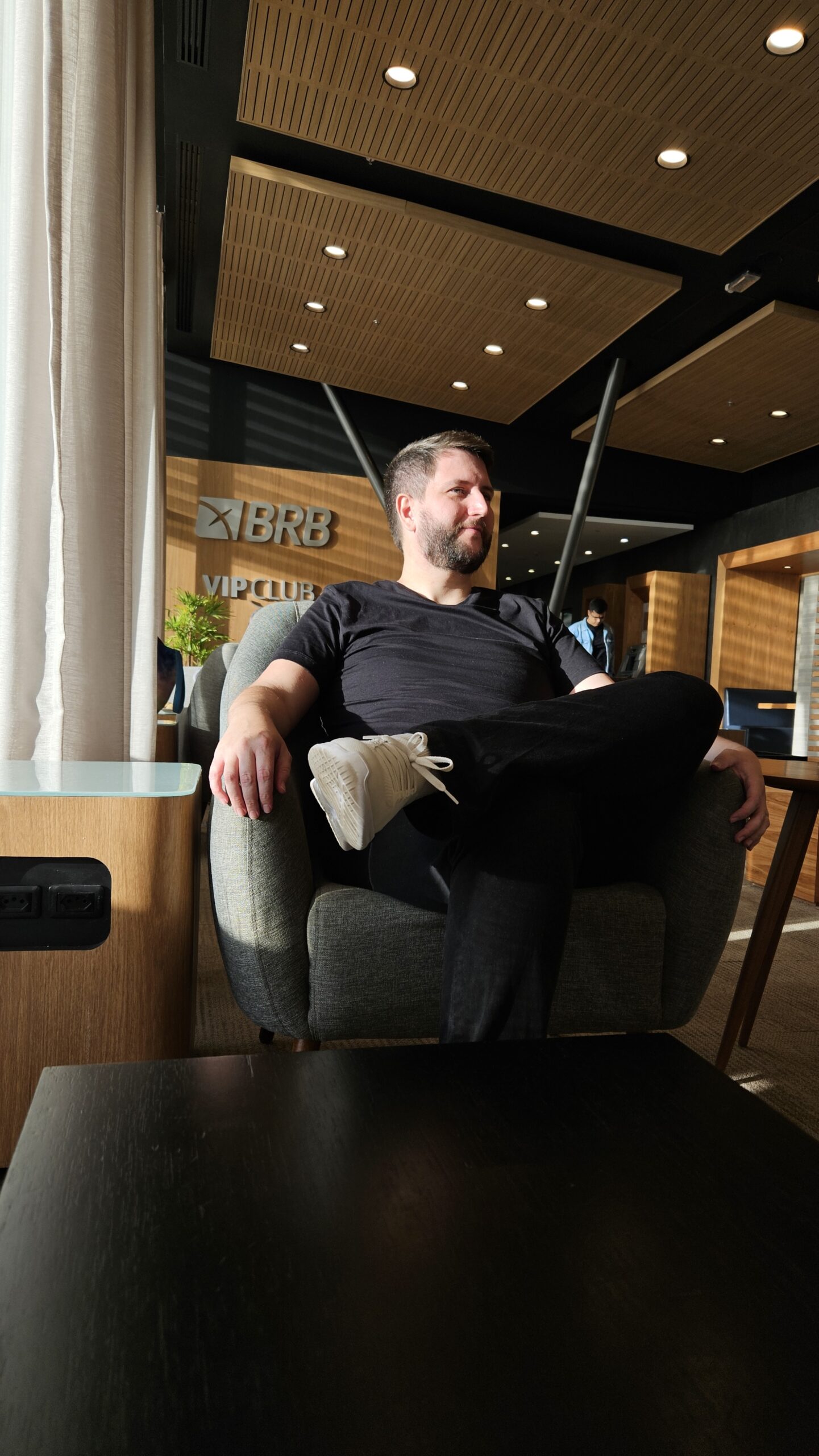 Rudi sentado no sofá da Sala VIP BRB de Brasília, com a placa BRB VIP Club ao fundo