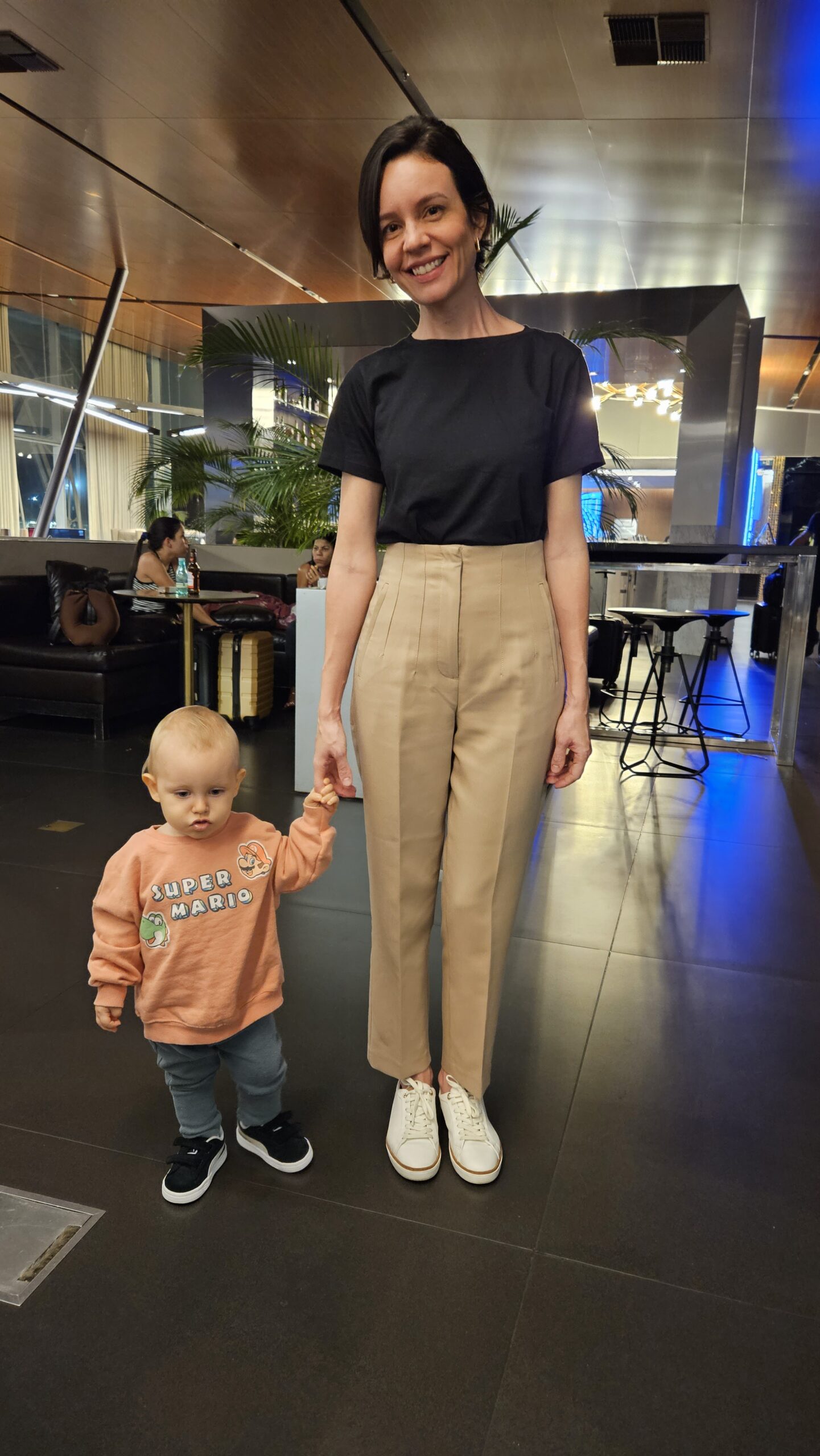 Carol e Samuel caminhando pela Sala VIP Club do Aeroporto de Brasília durante visita noturna, com iluminação azul do bar ao fundo