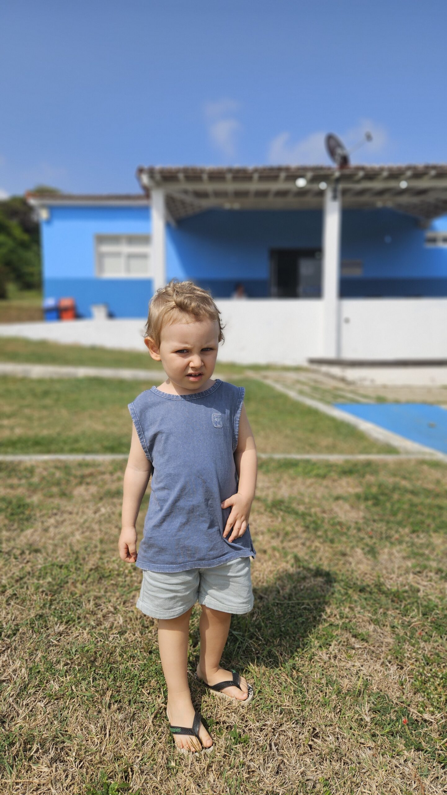 Criança pequena em pé na grama em Fernando de Noronha, em frente a um prédio azul e branco
